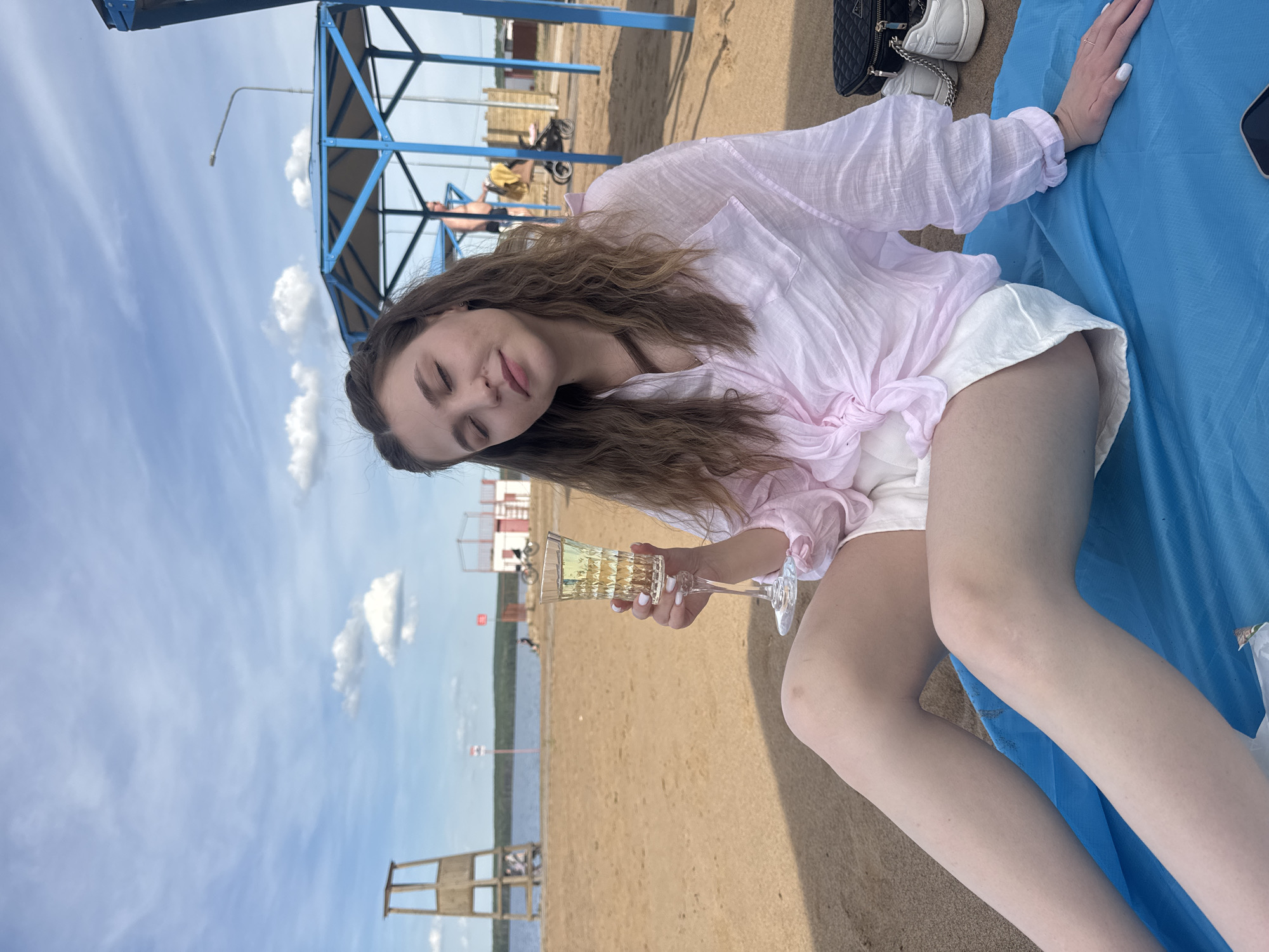 A woman is sitting on a blue blanket on the sand. She is wearing a light pink shirt tied at the waist and white shorts. She is holding a glass of yellow liquid in her right hand. There is a white purse and shoes next to her on the blanket. In the background, there is a sandy beach, a body of water, and a blue shelter structure. There is also a lifeguard stand in the distance. This location is in the city of Chaikovsky, Russia.