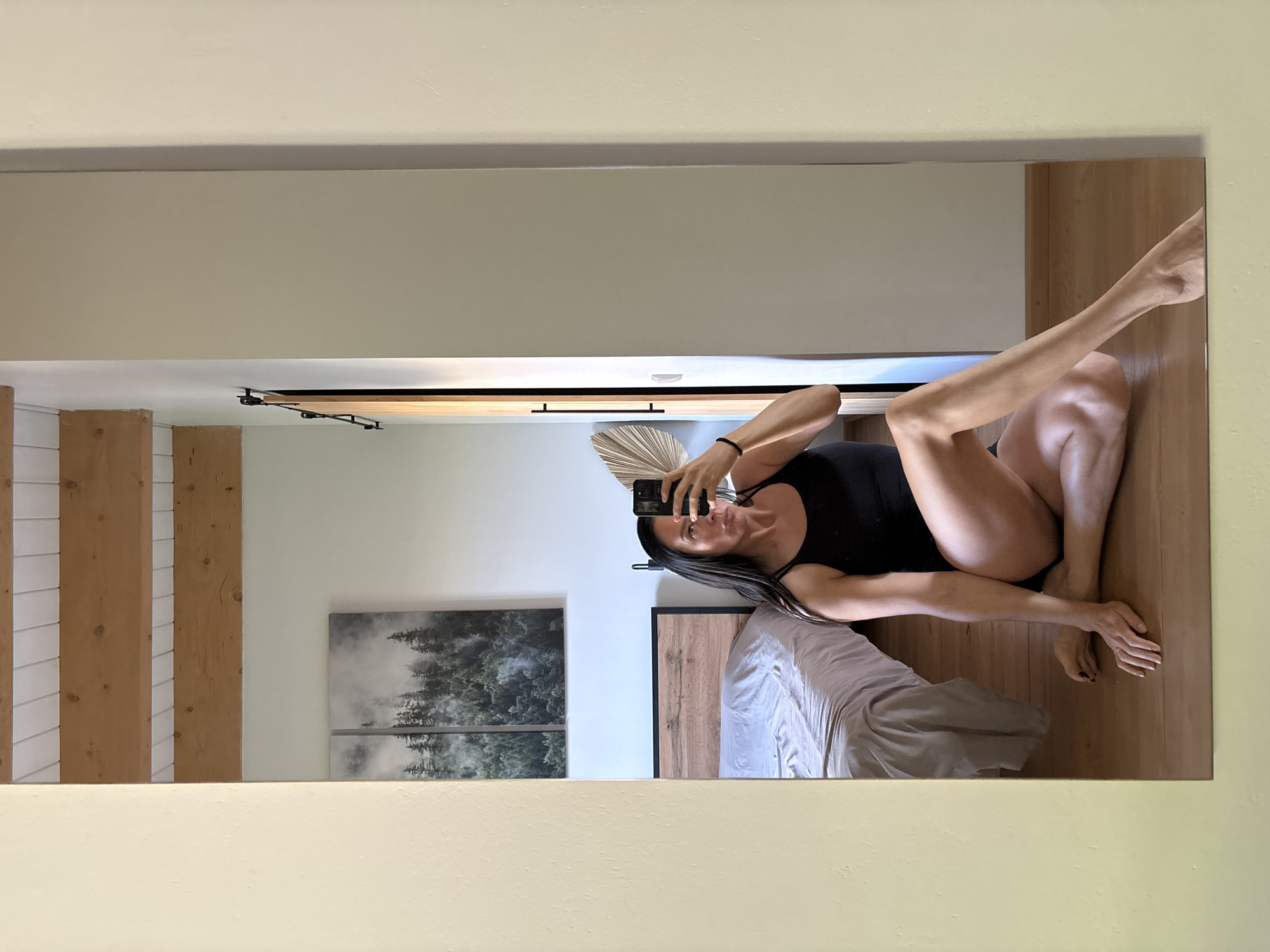 A full-length mirror reflects a woman sitting on a wooden floor in a bedroom. She is wearing a black tank top and black bottoms, and is holding a smartphone to take the picture. The bedroom has a bed with a grey comforter and a wooden headboard. Above the bed, there are two framed prints of a foggy forest. The ceiling has exposed wooden beams. The room is brightly lit, and the mirror reflects the entire scene. The location is not specified to be in Chaikovsky, Russia.