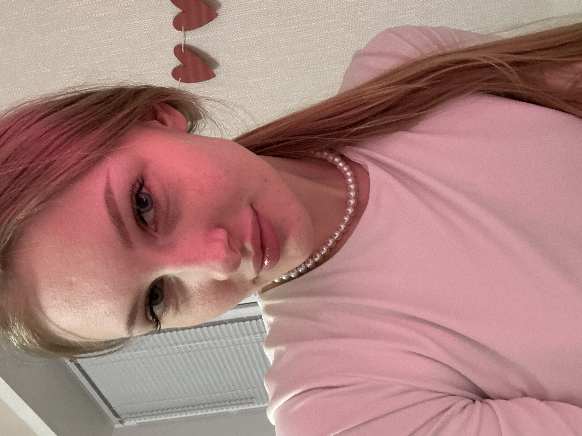 A close-up portrait captures a young woman, likely in her late teens or early twenties, positioned upside down from a low-angle perspective. She has long, light brown or reddish-blonde hair that falls over her shoulders and partially obscures her ear. Her eyes, appearing blue or gray, look directly at the viewer, framed by visible eyelashes. Her lips are full and glossy, suggesting a neutral or subtly smiling expression. She wears a string of pearls around her neck, visible over a light pink or white long-sleeved top or dress, which shows soft folds.
The lighting in the image casts a strong pinkish or reddish glow on her face and hair, creating a distinct aesthetic. In the background, a textured white ceiling or wall is visible, from which two red heart-shaped decorations hang. Below her head, a white gridded structure, possibly a ceiling vent or light fixture, is partially seen, indicating an indoor setting. The overall mood is calm and personal, suggesting a casual photograph taken in a private space, potentially in Perm, Russia. No visible text is present in the image.