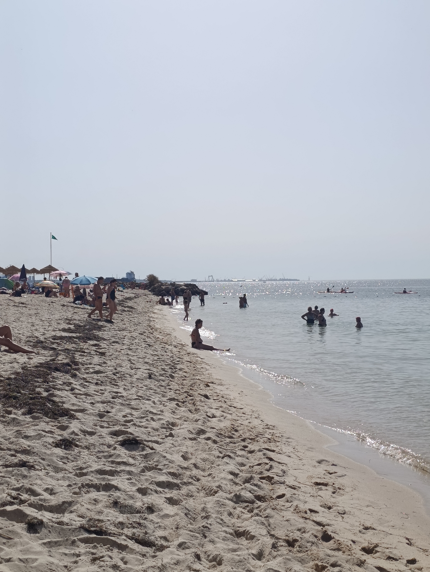 This is a photograph of a sandy beach on a sunny day. The beach is populated with many people, some sunbathing, some walking, and others in the water. There are several beach umbrellas and a green flag visible on the beach. In the background, there is a body of water with several boats and kayaks, and a distant industrial skyline. The scene appears to be from the city of Setúbal, Portugal, likely Praia da Saúde or a nearby beach. The water is calm and clear, with sunlight glinting off the surface. The sand is light brown and has some scattered seaweed. The sky is a pale blue with a few faint clouds.