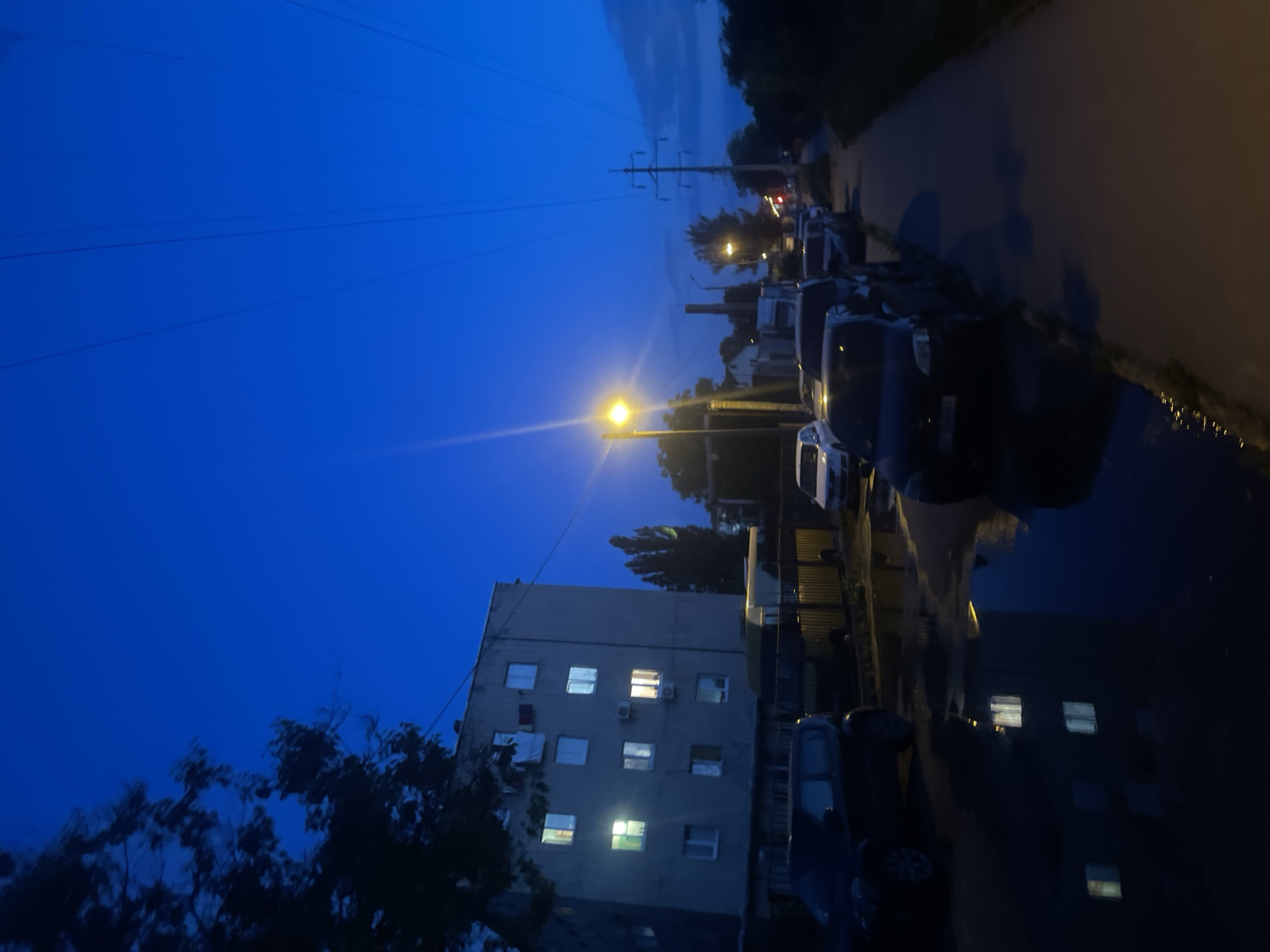 The image depicts a street scene at dusk or night, with a deep blue sky overhead. A multi-story apartment building with illuminated windows is visible on the left. Several cars are parked along the street and on the sidewalk, some of which are reflected in puddles on the wet ground. A street lamp is lit on the right side of the image, illuminating the walkway. Power lines and poles are visible against the sky, suggesting an urban or suburban environment. Trees and a chimney stack are also present in the background. The overall atmosphere is dark and quiet, with artificial lights providing the primary illumination.