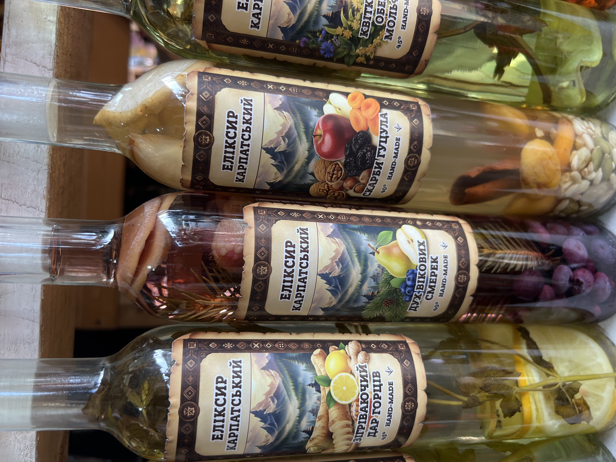 The image shows a close-up of four glass bottles containing various infusions, displayed on a wooden shelf. These bottles, likely for sale, have decorative labels with Cyrillic text and imagery of mountains and natural ingredients. The leftmost bottle contains sliced lemons and herbs. The second bottle from the left has a dark red liquid with what appears to be dried fruits. The third bottle contains dried fruits like apricots and prunes, along with nuts and an apple. The rightmost bottle appears to contain herbs and some yellow fruit slices. The bottles are arranged in a row, and the background suggests an indoor setting, possibly a shop or market stall. Given the labels and context, this scene is consistent with the artisanal food and souvenir shops found in Yaremche, Ukraine, a popular tourist destination in the Carpathian Mountains. There are no people visible in the image.