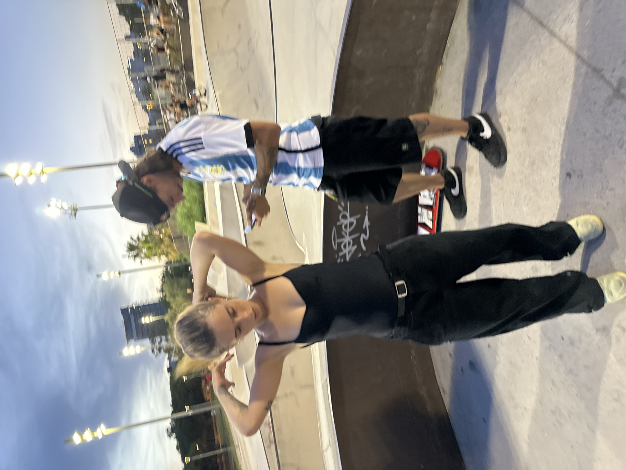 The image captures a dimly lit outdoor skate park at dusk, with artificial lights illuminating the scene. Two individuals are present. In the foreground, a young woman with blonde hair pulled back is adjusting her hair with both hands, wearing a black tank top and black pants with a belt. To her right, a man wearing a black baseball cap, an Argentina soccer jersey with blue and white stripes, and black shorts is looking down at his mobile phone. A skateboard rests on the ground near his feet. The skate park features concrete ramps and walls, with graffiti visible on some surfaces. In the background, city buildings and palm trees are visible under a cloudy sky. The overall atmosphere is casual and relaxed.