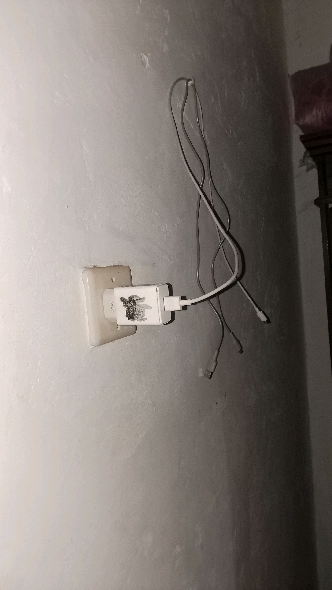 A close-up, eye-level, indoor shot shows a white electrical outlet and a white Oppo brand charging brick plugged into it. The charging brick has scorch marks on its surface, indicating damage. Two charging cables are connected to the brick; one is plugged into it, and the other hangs loosely. The wall behind the outlet and brick is textured and painted a light color. In the background, to the right, a dark, blurred object with a reddish-pink covering is visible, possibly furniture. The lighting suggests it is nighttime or the room is dimly lit. The scene is located on a wall in Mulyo Haji, Indonesia.