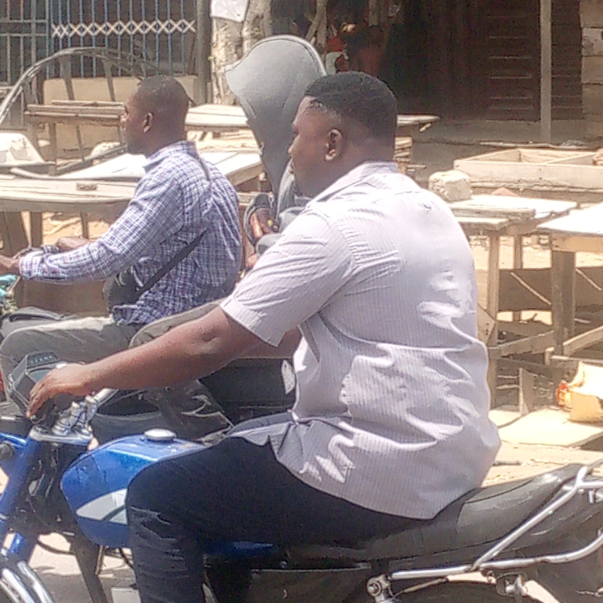 Two men are on a blue motorcycle, moving through an outdoor area. The motorcycle is the primary subject. The man closest to the camera is wearing a light-colored short-sleeved shirt and dark pants. The man further away is wearing a plaid shirt and light-colored pants. The location appears to be Adankolo, Nigeria. Background details include tables and structures that suggest a market or commercial area.