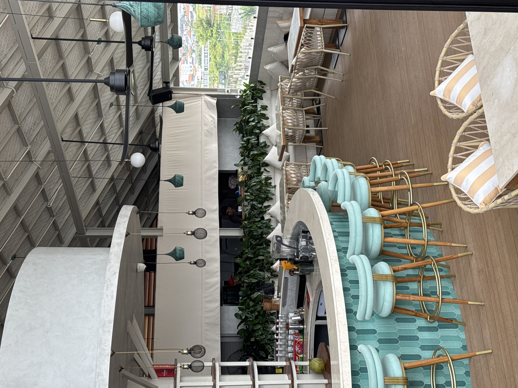 This is a wide shot of an open-air rooftop bar in North Kuta, Indonesia. The bar area features a curved, light-colored countertop with a teal tiled backsplash. Several teal bar stools with wooden legs and gold accents are positioned around the bar. Behind the bar, rows of bottles and bar equipment are visible. Green plants line the wall behind the bar.
The seating area in the background consists of light-colored banquette seating with white pillows, paired with rattan chairs and light wood tables. The floor is made of dark wood planks. The ceiling is a metal corrugated structure, with several black ceiling fans and pendant lights hanging down. A large window overlooks a city with buildings and greenery.
In the background, two men are partially visible behind the bar. One man wears a red cap and the other a dark shirt. Two wicker chairs with orange and white striped pillows are in the foreground, placed at a table. A coconut sits on the counter to the left of the bar. The overall ambiance is modern and relaxed, suggesting a dining or lounging establishment.