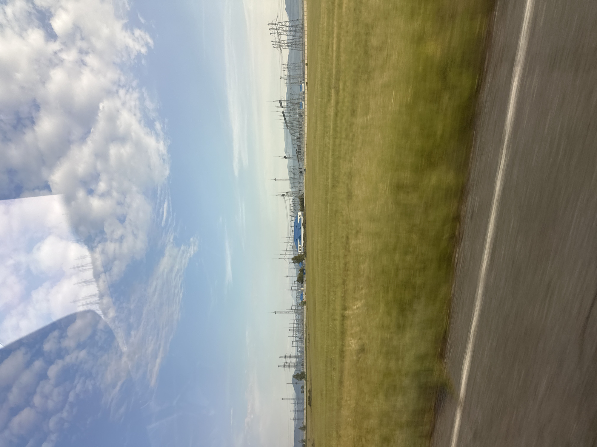 The image displays a scene captured from a moving vehicle, looking towards a vast, grassy field. A portion of the road is visible on the right side of the frame, with a distinct white line marking its edge. Beyond the field, a power substation with numerous tall metal towers and electrical wires stretches across the landscape. In the distance, mountains can be seen, partially obscured by the power infrastructure. The sky is a clear blue with scattered white clouds, suggesting fair weather. The lighting indicates it is daytime. The blurriness of the image suggests the photo was taken while in motion, likely from a car or train. There are no people or animals visible in the frame. The overall impression is of an open, rural or semi-industrial landscape under a bright sky. There is no visible text in the image.