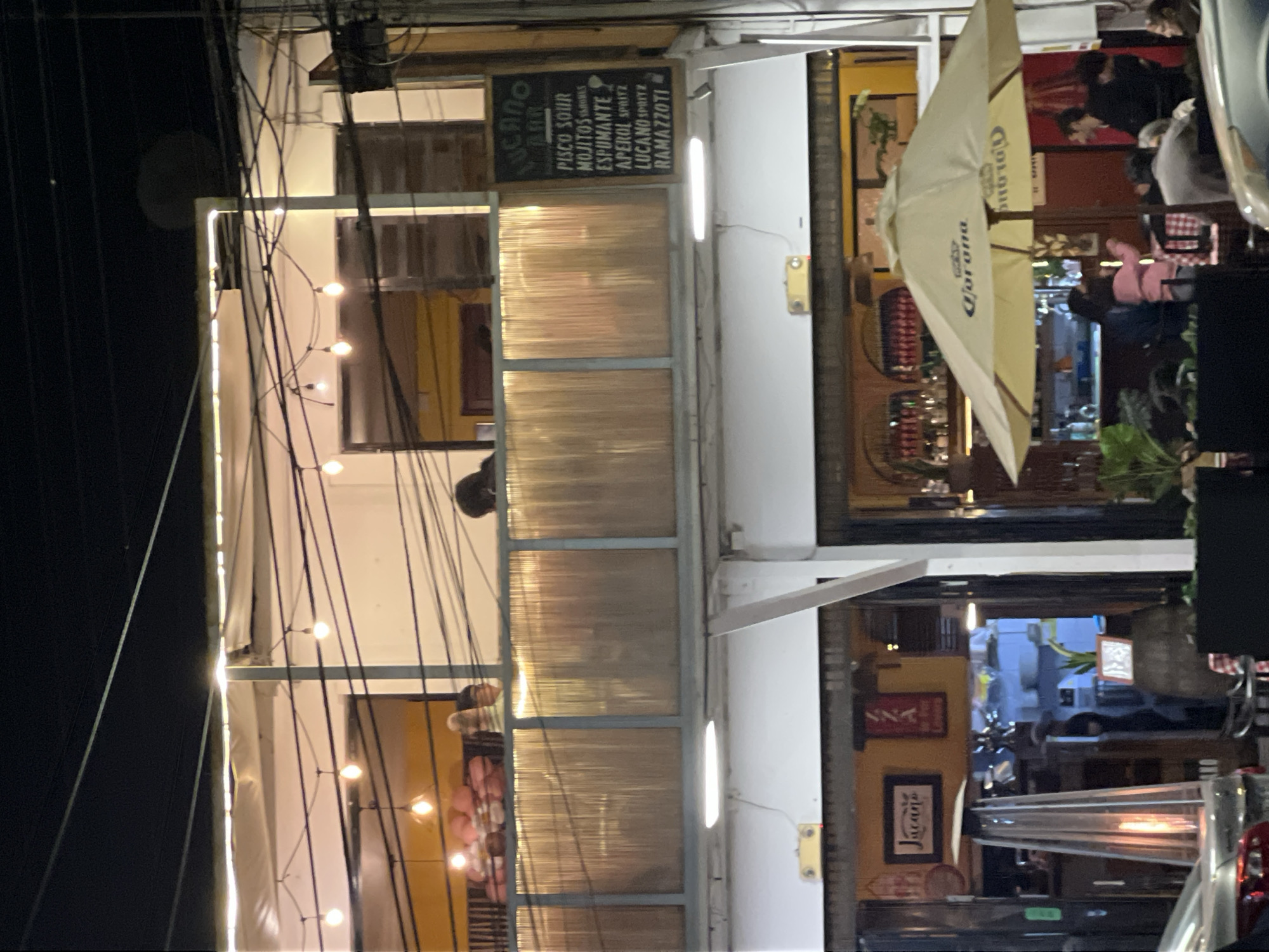 This appears to be an outdoor shot of the Lucano Bar in Arica, Chile, taken at night. Patrons are seated inside the bar. A Corona umbrella is positioned on the patio. The bar's sign lists drinks, including Pisco Sour, Mojitos, Espumante, Aperol Spritz, Lucano Spritz, and Ramazzotti. String lights are visible.