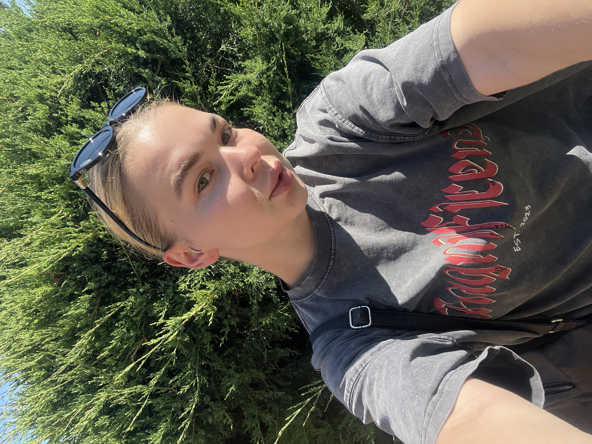 The media file is a vertical, close-up selfie of a young woman with blonde hair and light skin. She is wearing a dark gray t-shirt with a distressed, red graphic design on the front that includes stylized text and "EST. 2023". Black sunglasses are perched on top of her head. A black strap, possibly from a bag, is visible across her chest.
The woman is positioned in front of a dense, green evergreen bush, likely a juniper, suggesting an outdoor setting. The sunlight indicates it is daytime, and the bright lighting implies fair weather. Her expression is one of slight contemplation or a casual pout. She has a small, delicate nose ring in her left nostril and a crescent-shaped earring on her right ear.
The background consists entirely of the green foliage of the bush and a bright blue sky peeking through in the upper left corner. The overall impression is of a relaxed, summery outdoor moment.
There is visible text on the t-shirt: a large, stylized red graphic followed by "EST. 2023".