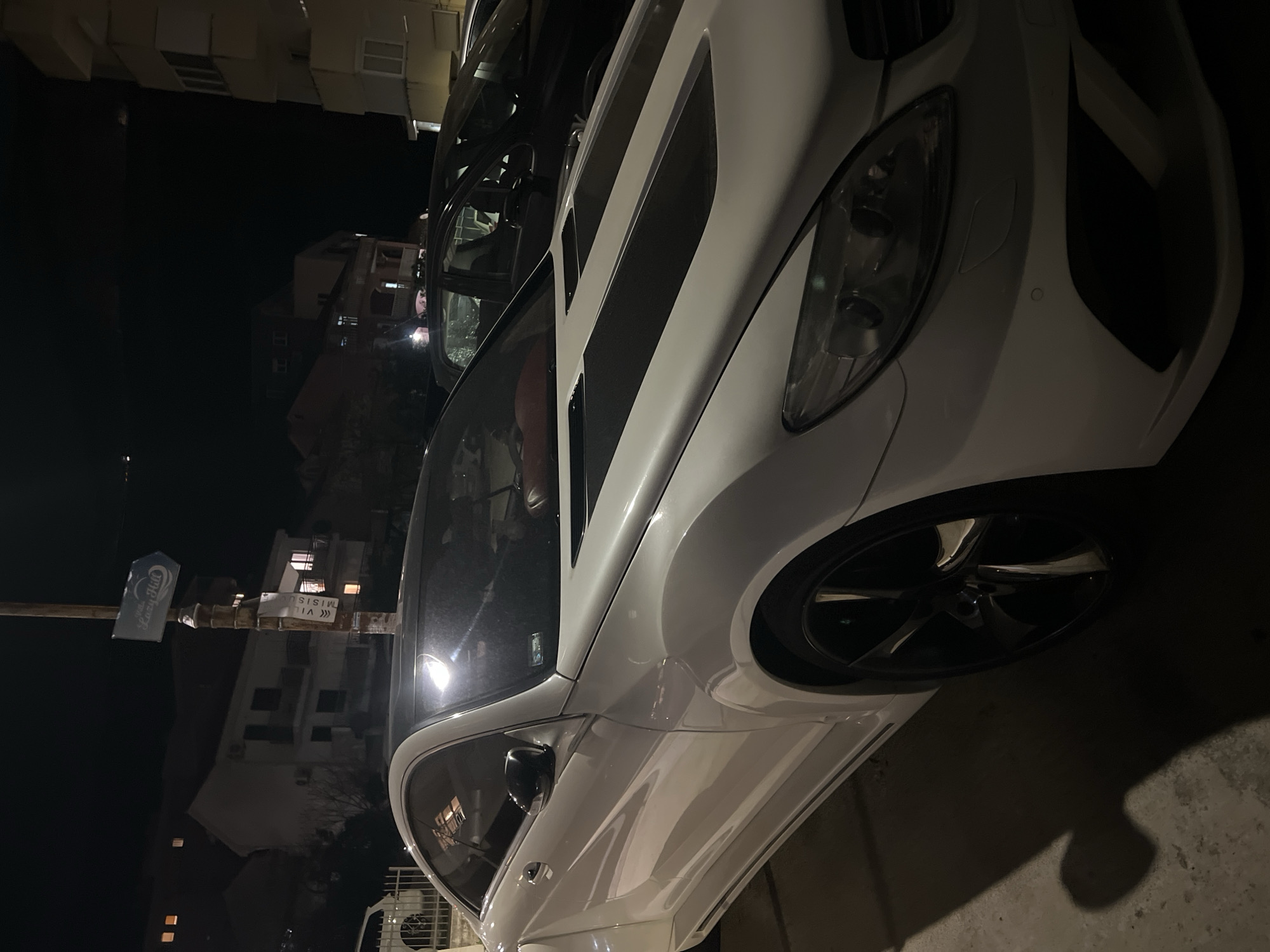 The image shows a white Mercedes-Benz parked at night. The car's front end is prominently visible, including the headlight, wheel, and part of the hood. The background shows buildings, suggesting an urban environment. A street sign with text "Lazy Hut" and "VIZI" is visible nearby. Based on the general appearance and surroundings, it could be in the city of Budva, Montenegro. No people are visible.