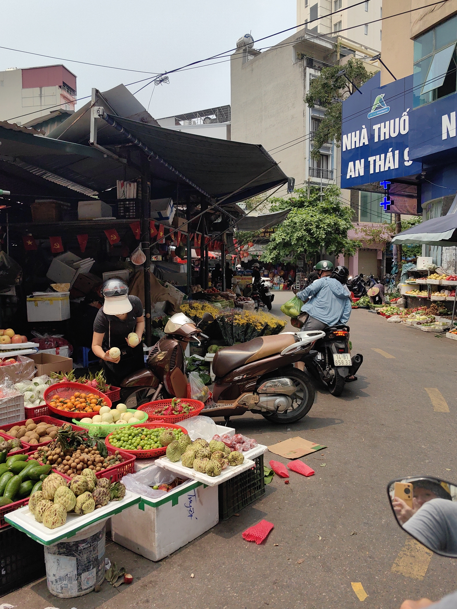 Here's a detailed description of the image:
**Location:** The image appears to be taken at a street market located in Hong Gai Ward, Vietnam. This is inferred from the overall visual style, language on the signs, and general aesthetic.
**Overview:** The scene depicts a bustling street market filled with vendors and customers. The foreground shows a variety of tropical fruits arranged for sale, with people moving around.
**People:** A woman wearing a face shield and helmet is seen holding fruits near a fruit stand. Another person on a motorbike is seen further up the street, wearing a helmet and jacket. The reflection in the side mirror shows a person holding a phone.
**Subjects & Objects:**
* Various fruits are on display in baskets and containers, including dragon fruit, mangoes, avocados, and other local varieties.
* Motorbikes are parked along the side of the street, likely belonging to shoppers or vendors.
* A pharmacy sign "NHÀ THUỐC AN THÁI" is visible on a building in the background.
* Overhead, there are awnings or coverings providing shade for the market stalls.
**Actions & Interactions:** The woman is selecting or examining fruits. The person on the motorbike seems to be driving away. The person in the reflection is taking a photograph.
**Environment:** The setting is an urban street. Buildings are seen in the background, along with overhead power lines. The street appears to be busy with activity.