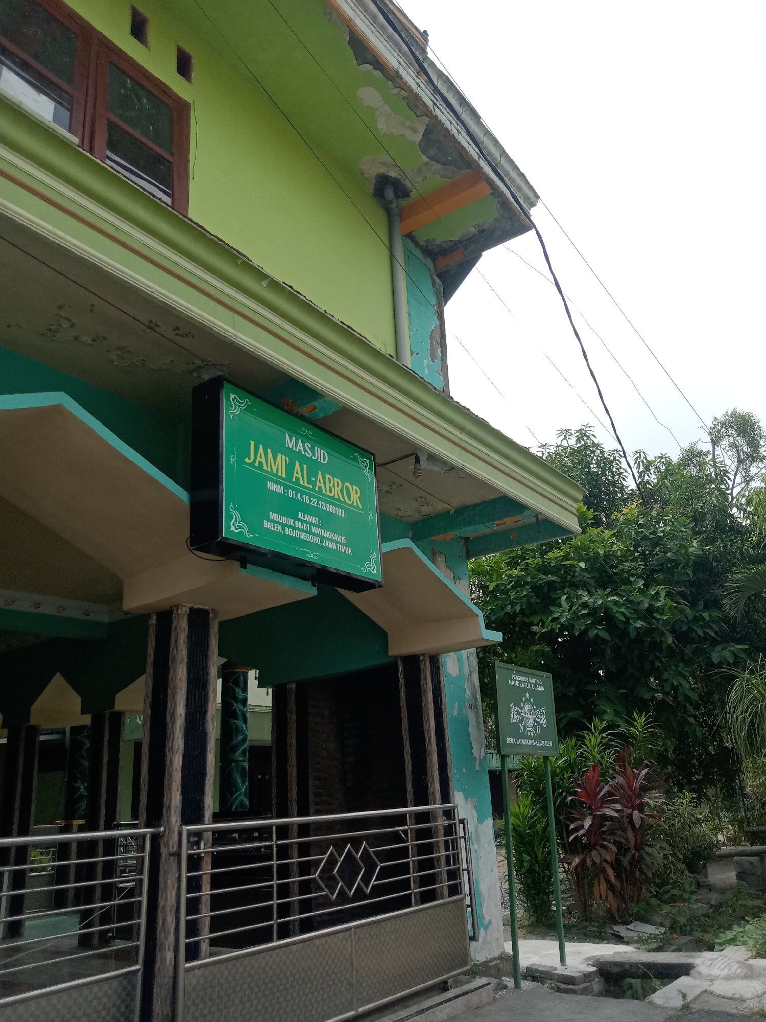 The image shows the exterior of a mosque named Jami' Al-Abror, located in Mayangkawis, Bojonegoro, Jawa Timur, Indonesia. The mosque building has a light green exterior with some signs of wear and tear, particularly on the upper facade. A prominent green sign with white lettering identifies the mosque and displays its NINM (Nomor Induk Nasional Madrasah) and address. Below the sign, there are pillars and a metal gate. To the right of the mosque, a green sign with text and a logo indicates "PENGURUS RANTING NAHDLATUL ULAMA" in Desa Mayangkawis. Lush green trees and foliage are visible behind the signs and to the right of the building. The sky is overcast.