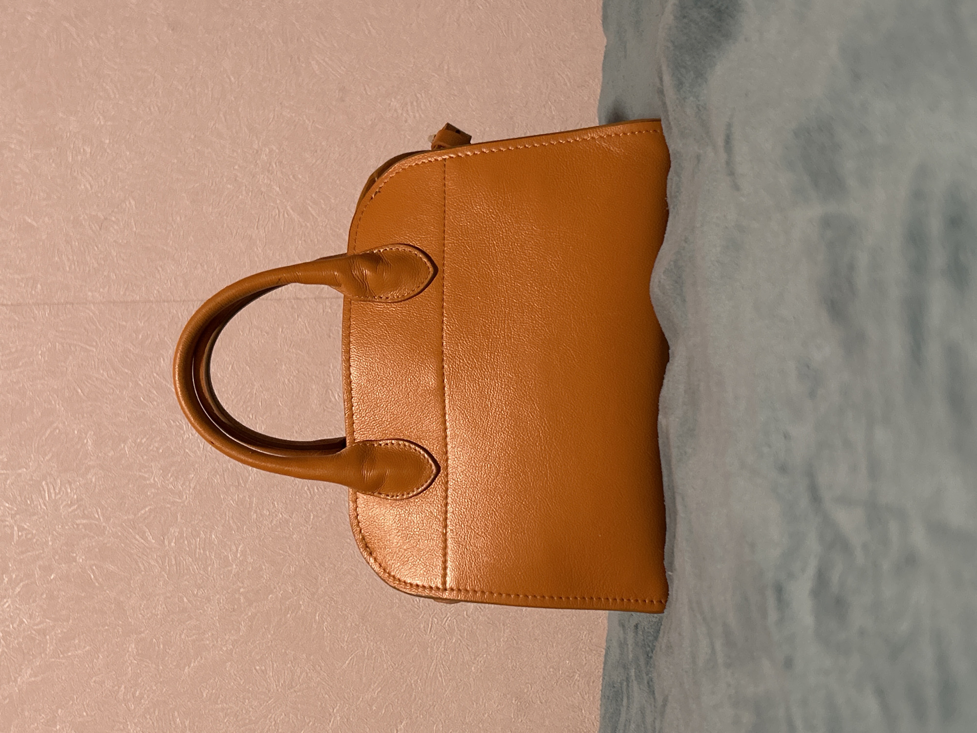 A medium shot, eye-level photograph shows a tan leather handbag on a blue fluffy blanket. The handbag has two rounded handles and a zip closure. The background is a light pink textured wall. The image is lit with warm, artificial light. The photograph was taken in Moscow, Russia.