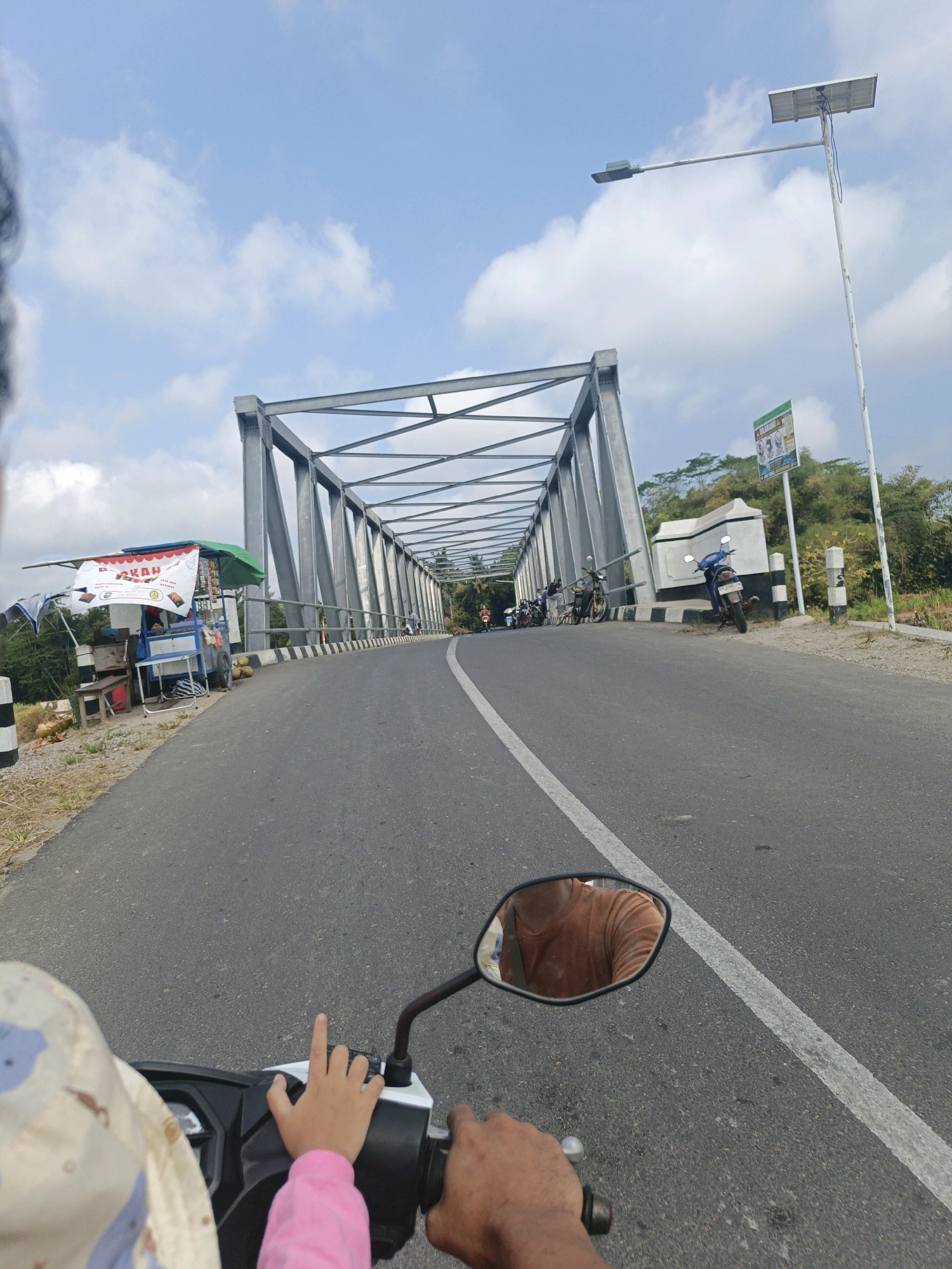 The image shows a view from a motorcycle moving along a road that crosses a metal truss bridge. In the foreground, a person's hand is visible on the handlebar, and a child's hand is on the dashboard. The child is wearing a pink long-sleeved shirt. The motorcycle's side mirror reflects the driver, who is wearing an orange t-shirt.
The bridge spans over a natural landscape with trees and vegetation visible on the sides. Along the roadside before the bridge, there is a small food stall with a green umbrella and various items for sale, including what appear to be coconuts. Further along the road, several motorcycles are parked, and people are gathered. A lamppost with a solar panel is positioned near the bridge entrance, and a sign with multiple warnings is mounted on it. The sky is blue with scattered white clouds. The road has a white dividing line. This scene is located in Wetonkulon, Indonesia, as indicated by the presence of local signage and the architectural style of the bridge and roadside stalls.