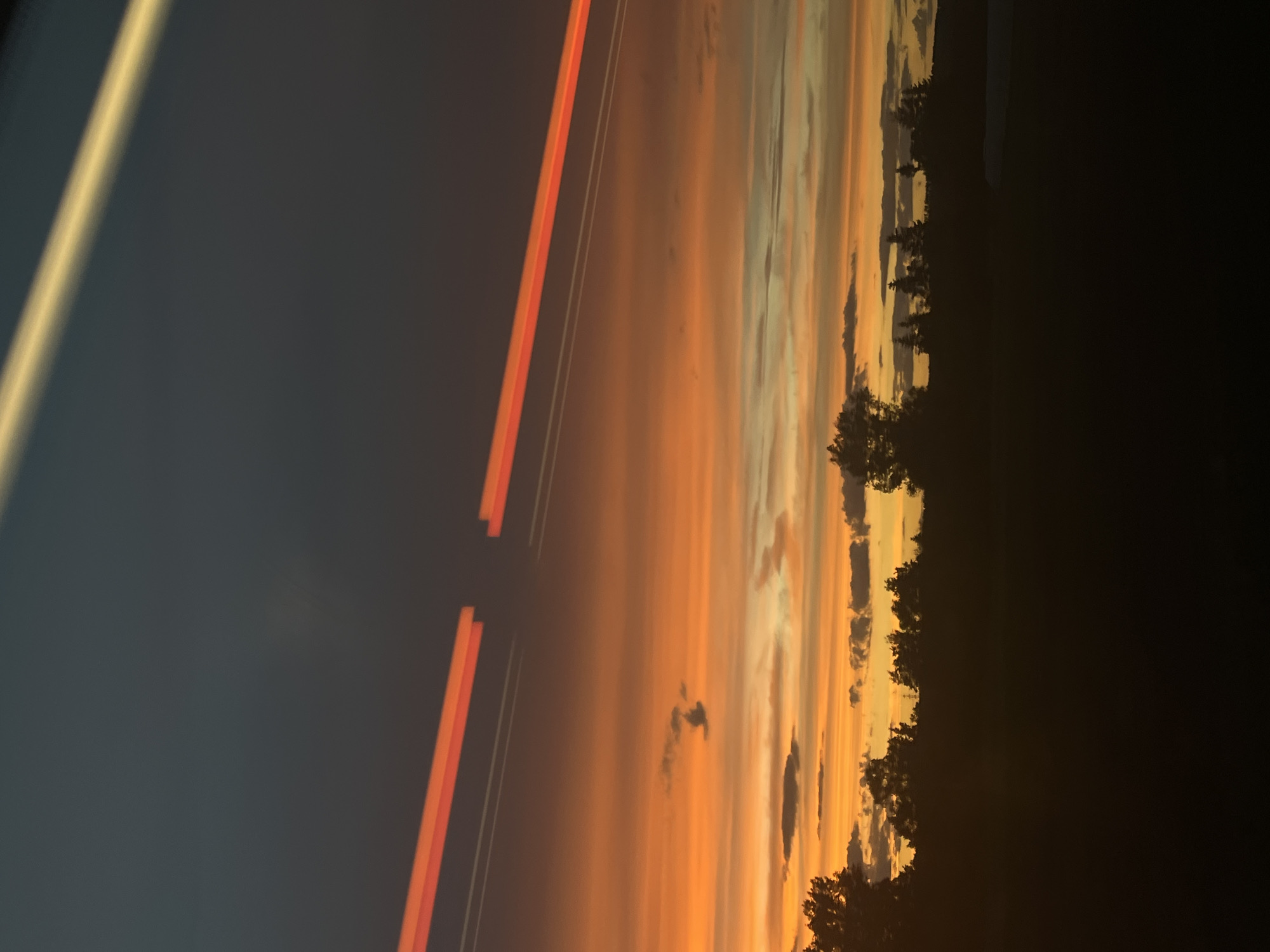 A sunset is captured over a silhouetted landscape of trees and fields. The sky is a vibrant gradient of orange, yellow, and dark blue, with streaks of light that appear to be from vehicle headlights or contrails. The clouds are scattered throughout the colorful sky, some appearing dark and others illuminated by the setting sun. The lower portion of the image is dominated by a dark silhouette of trees and vegetation, suggesting a rural or suburban area. The scene is likely captured from a moving vehicle due to the light streaks. The image does not contain any identifiable landmarks or features that would place it specifically in Baiersdorf, Germany.