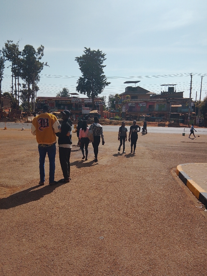A group of people walks across a dirt road in an outdoor area. Buildings with corrugated roofs and trees are in the background. A boy plays with a ball in the distance. The sun casts shadows of the walking individuals onto the reddish-brown road. A man wearing a yellow letterman jacket faces away from the camera. The sky is clear and blue.