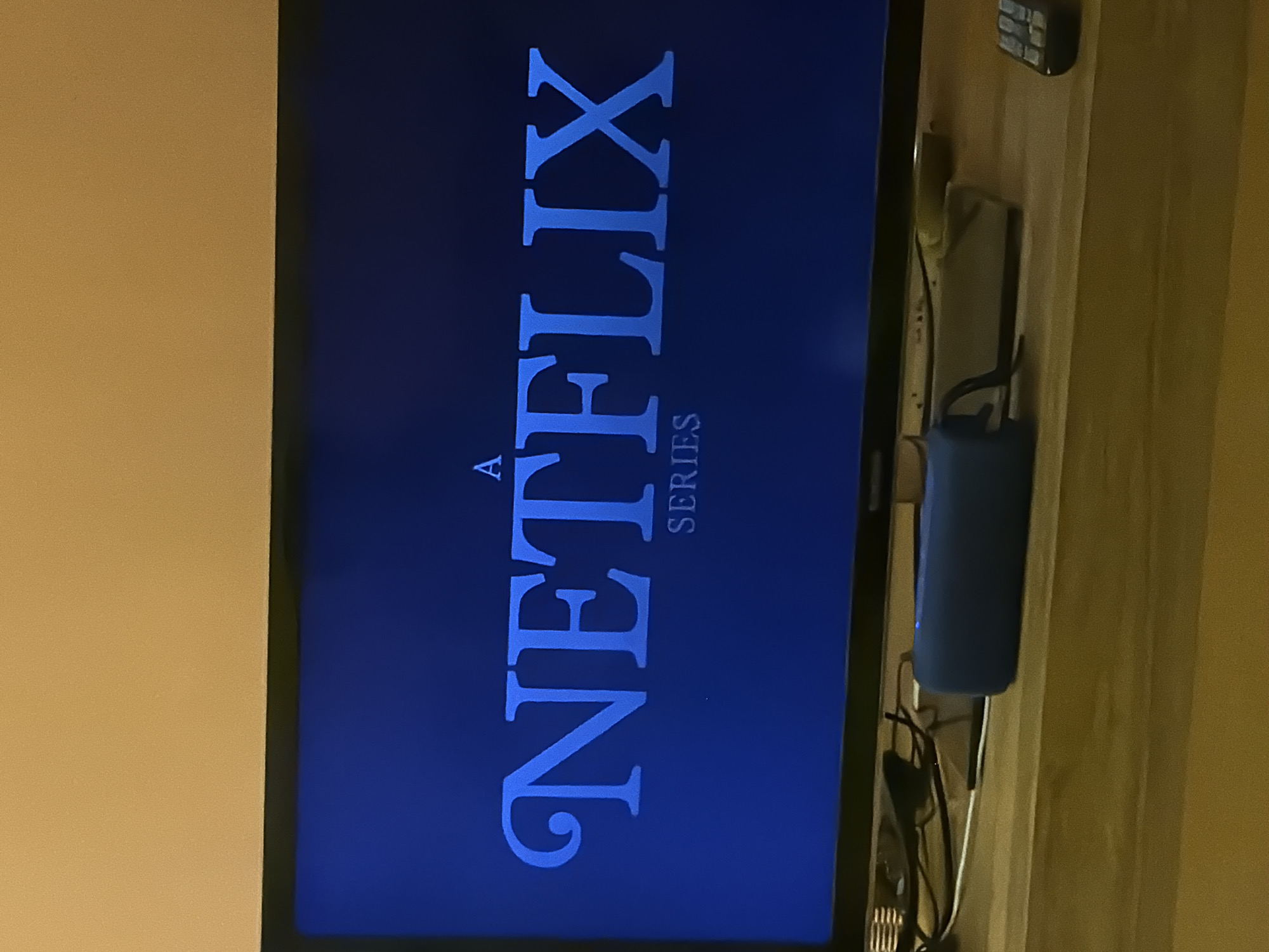 The image shows a television screen displaying the Netflix logo and the text "A SERIES" in blue on a dark blue background. The screen is situated on a wooden shelf alongside electronics, including a remote control. The wall adjacent to the television is painted a light beige or yellow color. There is no indication of the image being related to the city of Tel-Aviv, Israel.