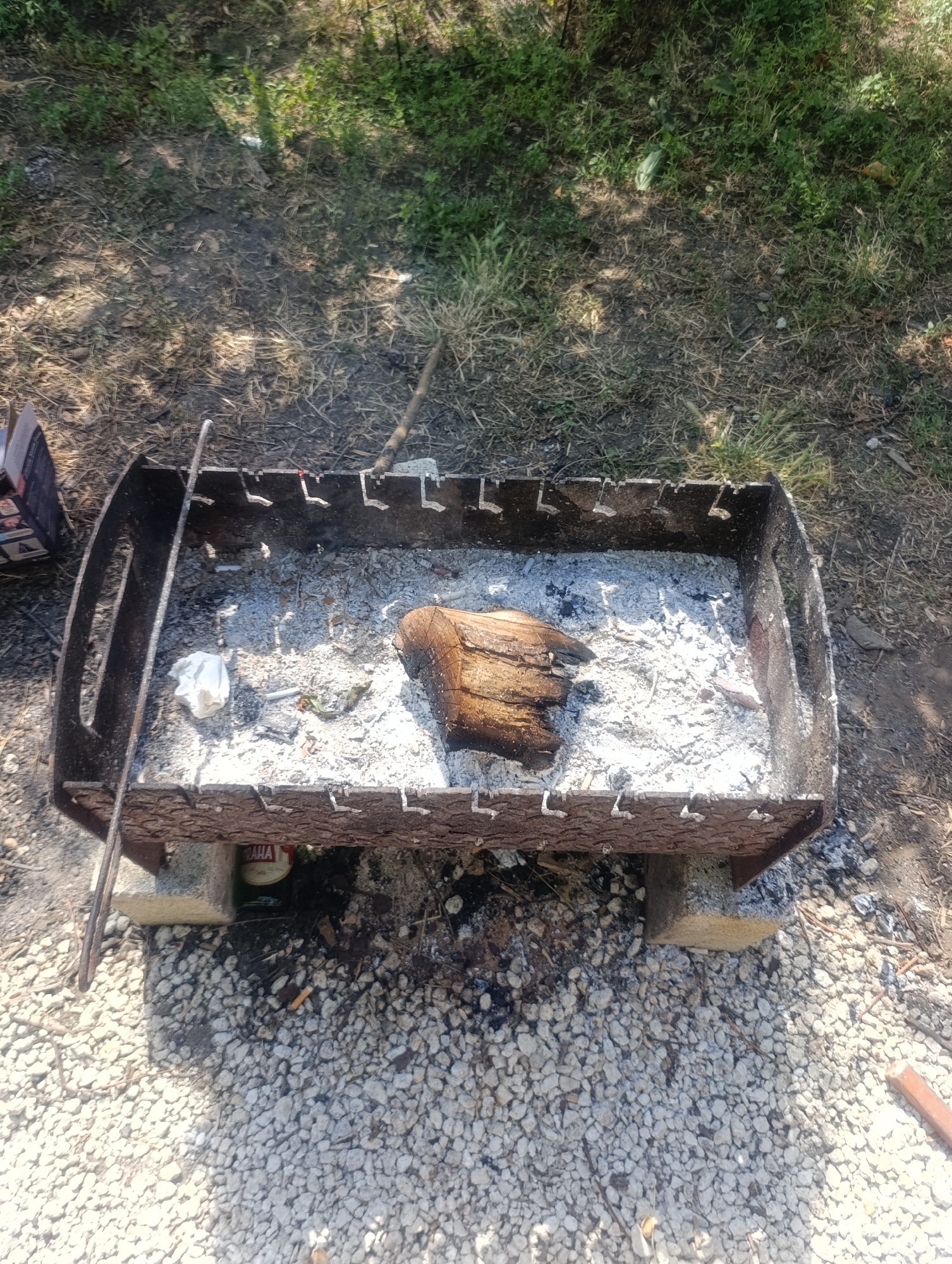 The image shows a portable metal barbecue grill filled with ash and a piece of wood, situated on a gravel surface. The grill is elevated by two concrete blocks. A green bottle with a label partially visible that says "BABA" is placed underneath the grill. A white crumpled paper and cigarette butts are also visible in the ash. The surrounding environment consists of dry grass and dirt, with shadows from trees indicating an outdoor setting. The scene appears to be in a natural or semi-natural area, likely a park or backyard, located in Dumeni, Moldova. There are no people visible in the image.