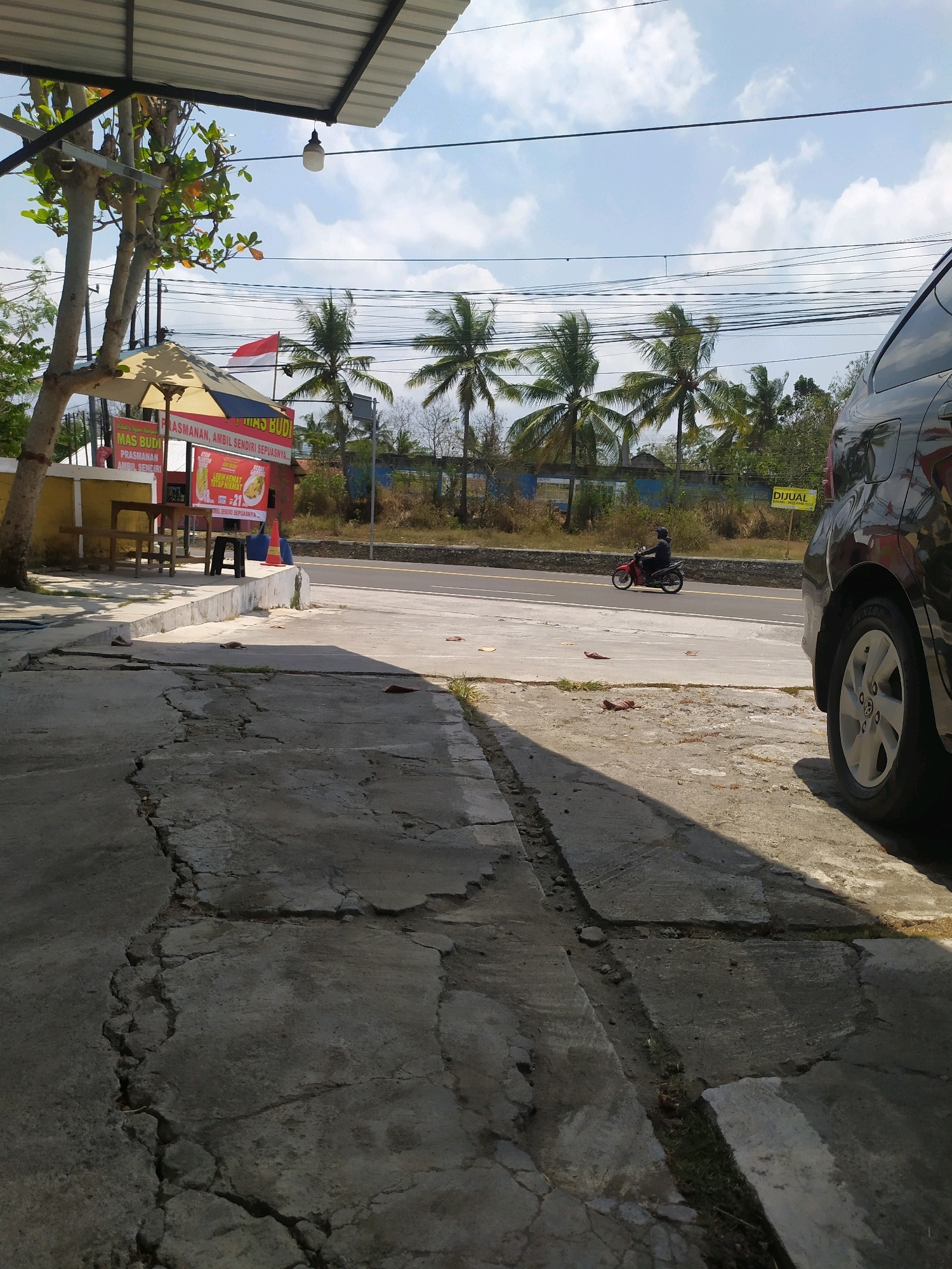 In Kedungsari, Indonesia, on a bright, sunny day, a roadside eatery stands partially visible on the left. A red and white Indonesian flag flutters from a pole. In front of the eatery, tables and chairs are set up, and a small, covered umbrella provides shade. A red and white sign with text is posted on a stand. The ground in front of the eatery is paved with cracked concrete. A black car is parked to the right of the scene, with its rear wheel and side visible. Across the street, a person on a red motorcycle rides down the road. Coconut trees and lush green foliage line the background, with a blue sky and scattered clouds above. A yellow sign is visible in the distance with the word "DIJUAL" (for sale) printed on it. The overall scene depicts a typical street view in a town or village in Indonesia.