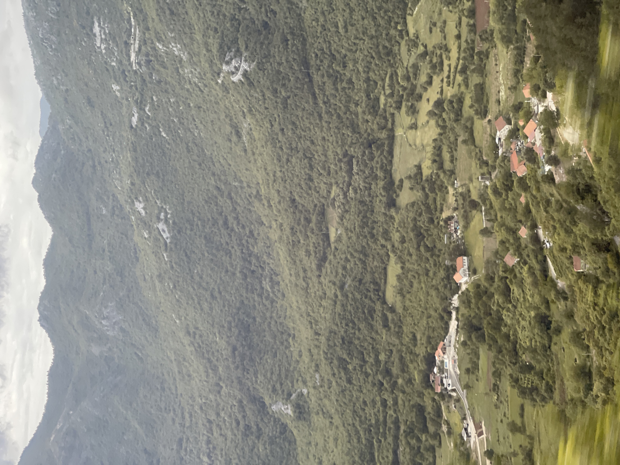 The image depicts a mountainous landscape, likely in the vicinity of Crna Greda, Montenegro. A steep, heavily forested mountain slope dominates the left side of the frame. The right side transitions into a more developed area with clusters of buildings, potentially residential, scattered along a narrow road. The buildings appear to have red-tiled roofs. The environment suggests a rural, mountainous setting.