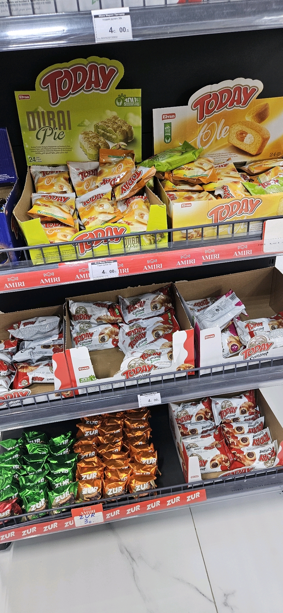 The image shows a retail shelf display in a grocery store in Dushanbe, Tajikistan. The shelves are stocked with various packaged snacks, primarily cookies and pastries from the brand "Today" and "Zur". Specifically, there are boxes of "Today Dubai Pie" and "Today Ole" cookies, along with packages of "Today Croissant Zerogram". Also visible are stacks of "Zur" branded individually wrapped snacks in green and orange packaging. A price tag for "Amiri Zur" at 30 Tajik Somoni is visible. The overall environment suggests a well-lit supermarket or convenience store aisle. There are no people or active interactions depicted. The display is neat and organized, with products arranged on metal shelves. The flooring appears to be tiled with a marble-like pattern.