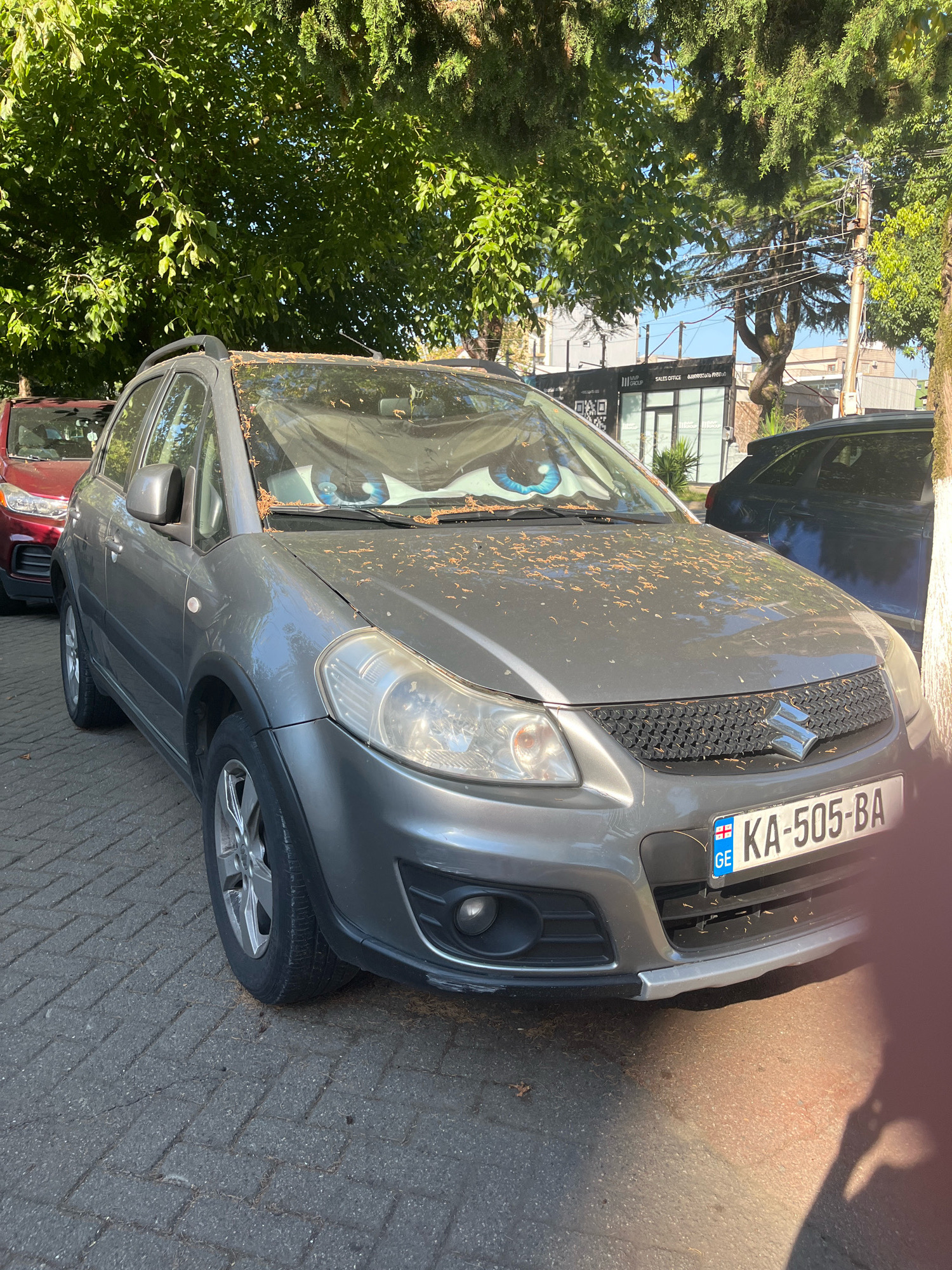 A parked gray Suzuki SX4 is visible from the front and side. The car has a license plate reading "KA-505-BA" and features a novelty windshield cover with large cartoon eyes. The vehicle is parked on a paved surface, possibly a sidewalk or street, with a red car visible behind it. Overhead, lush green trees provide shade, and power lines are strung between poles in the background. Buildings are also visible in the distance. The scene appears to be located in Kobuleti, Georgia, as indicated by the license plate and general environment.