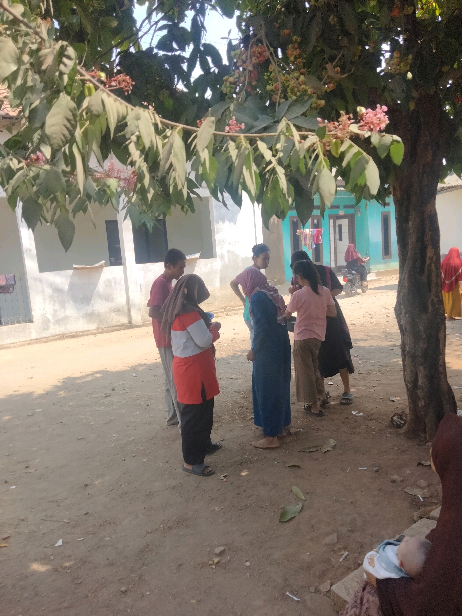 A group of people are gathered outdoors under a tree with pink blossoms in Suka Sari, Indonesia. The ground is dusty dirt. In the background, there are two single-story buildings. One is light colored with dark windows, and the other is turquoise with white trim. Several people are standing and talking. One woman, wearing a red and gray top and a headscarf, is holding a small blue bowl. Another woman in a blue dress and headscarf is facing away from the camera. A man in a red shirt is standing to the left. Further in the background, a person is riding a motorcycle in front of the turquoise building. To the right, a woman in a red headscarf and yellow skirt is visible, and further to the right, a person in a brown headscarf is holding a baby.