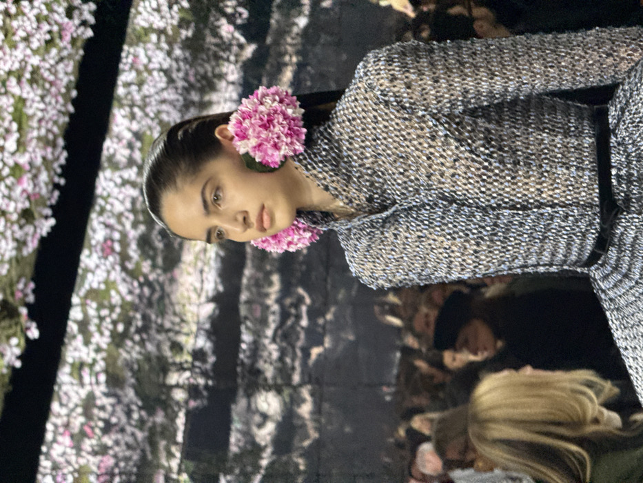 A model with slicked-back hair and bright pink floral earrings is captured on a runway. She is wearing a tweed jacket with a black belt. The backdrop features a blurred floral pattern, possibly reflecting onto a screen or wall. Other audience members are visible in the background, some with blonde hair. The lighting is bright, highlighting the details of the fabric and the model's makeup.