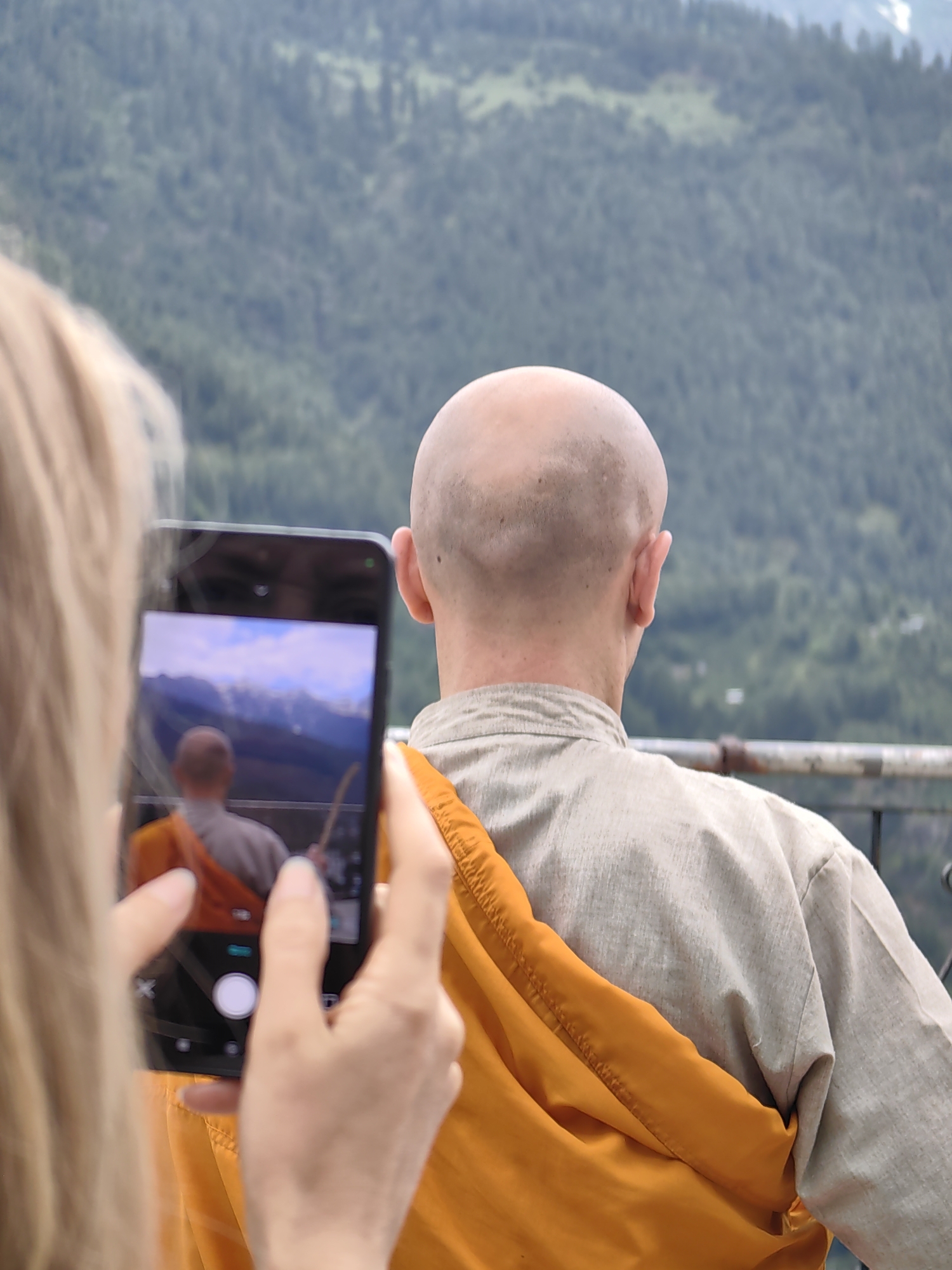 In the city of Vashisht, India, a person with blonde hair is taking a photo with their smartphone of another individual. The subject of the photograph, facing away from the camera, is a bald person wearing a light-colored shirt and an orange robe draped over their shoulders. The background is a lush, green, mountainous landscape with trees and a hazy sky. The photo on the smartphone screen shows a similar mountain scene and the back of the bald person, suggesting a selfie or a photo taken by someone else and viewed on the phone.