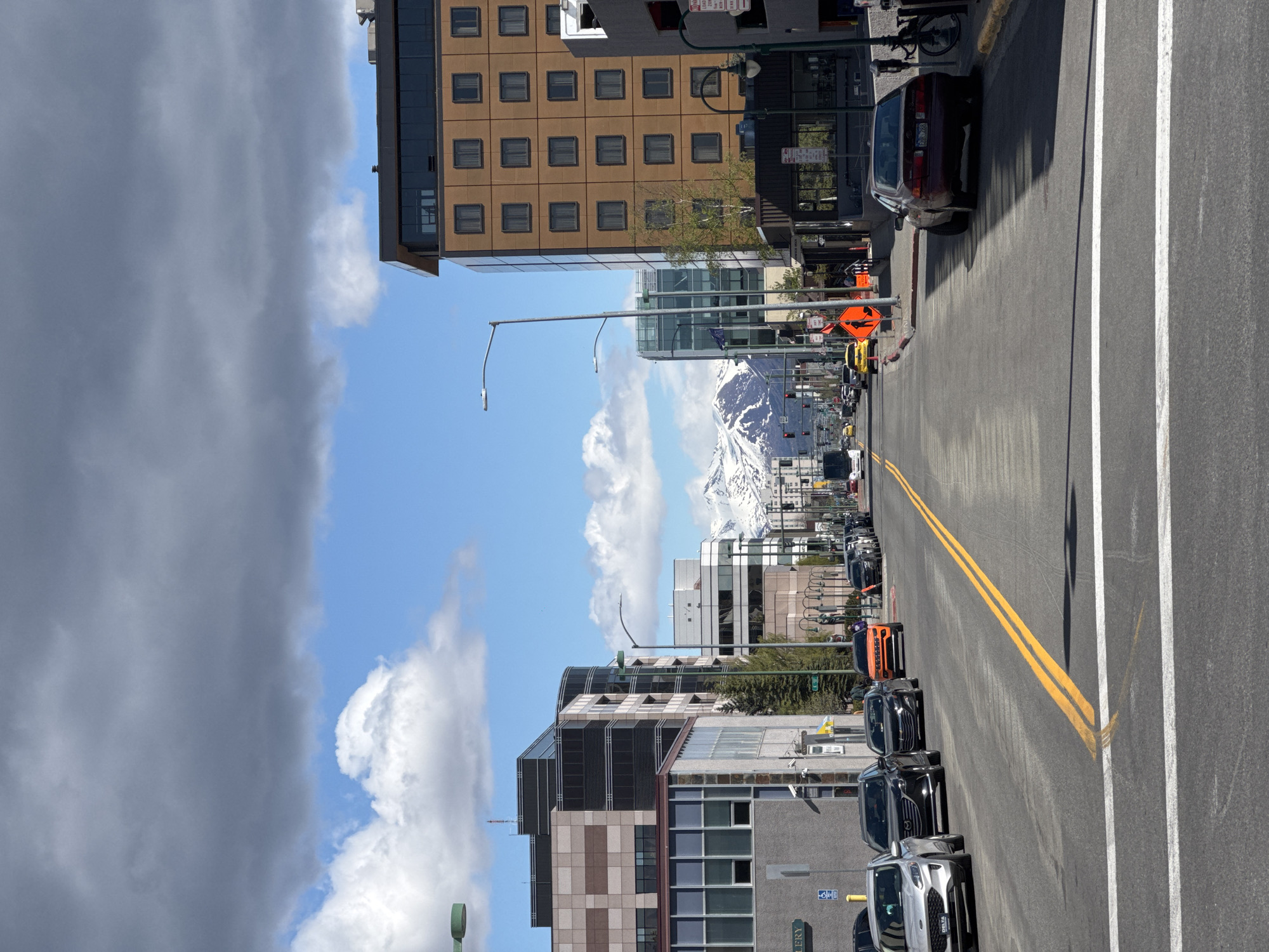 The image captures a street view in Anchorage, Alaska, United States. The scene is taken from the side of the road, showing cars parked along the curb. An orange car and several other vehicles are visible, parked in a line. The road features double yellow lines marking the center and a white line along the edge. The background includes buildings of varying heights and architectural styles. A mountain range, partially obscured by clouds, can be seen in the distance under a clear, blue sky. The sky also has some gray clouds. Overall, the image presents an urban setting with a mountainous backdrop.