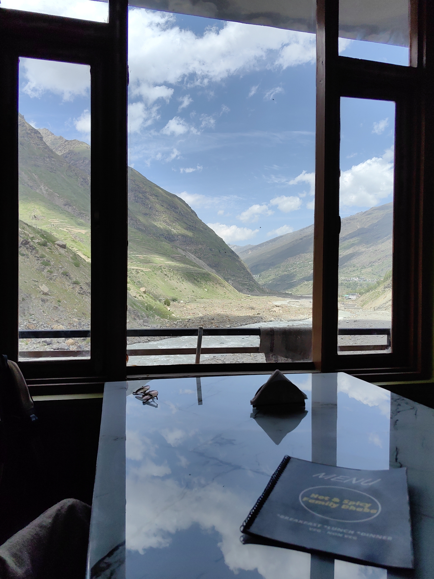 The image depicts an interior view from a room looking out a large window at a mountainous landscape in Sissu, India. In the foreground, a reflective marble-topped table holds a napkin holder and a menu. The menu, titled "MENU," appears to be for a place called "Hot & Spicy Family Dhaba" and lists "BREAKFAST," "LUNCH," and "DINNER." Through the window, a river flows through a valley flanked by steep, green mountains, some with terraced farming. The sky is blue with scattered white clouds. The window frame is dark wood, dividing the view into three vertical panes. A blanket or towel hangs over a railing outside the window.