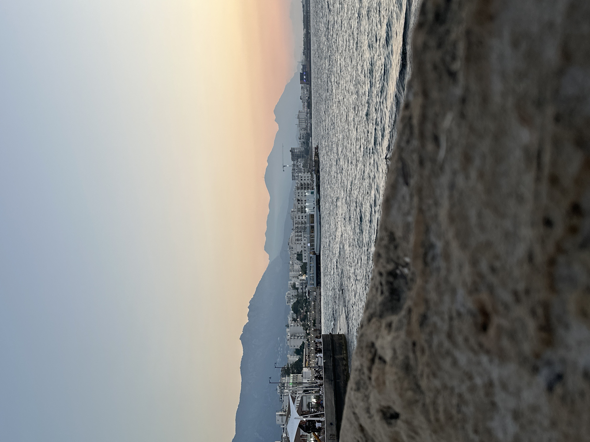 The image depicts a scenic view of Kyrenia, Northern Cyprus, at dusk. In the foreground, a rough, textured stone surface is visible, out of focus. Beyond this, a body of water, likely the Mediterranean Sea, is present, with gentle waves creating ripples on its surface. Along the shoreline, a promenade or pier is lined with buildings, some of which appear to be hotels or restaurants, with awnings providing shade. Several people are scattered along the promenade, engaged in various activities. In the distance, the silhouette of the Kyrenia mountain range is discernible against the soft, pastel hues of the twilight sky, transitioning from a pale blue at the top to warmer shades of orange and pink near the horizon. A few boats are visible in the water, and construction cranes hint at ongoing development along the coast. The overall atmosphere is peaceful and serene, capturing the tranquil beauty of a coastal city at sunset.