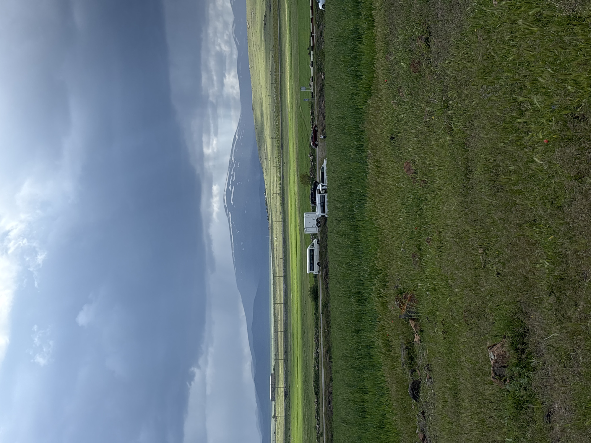 The image depicts a landscape scene in Tlik, Armenia. A mountain with snow patches is visible against a cloudy sky. In the foreground, there is a road with several parked white vans. Adjacent to the road are green fields. The overall impression is a rural or semi-rural environment.