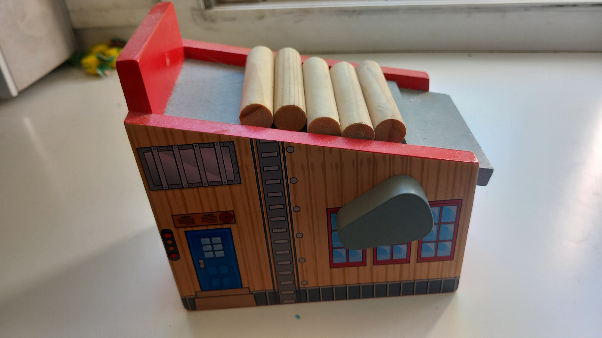 The image shows a wooden toy house with red trim and a gray roof. Five wooden dowels are placed horizontally on top of the roof. The front of the house is painted to resemble a building with a blue door, windows, and a ladder. There is a gray, oval-shaped object protruding from the side of the house, possibly a chimney or an air vent. In the background, out of focus, is a blurred yellow and green object, potentially another toy. The toy is placed on a white surface, possibly a table or shelf, with a bright, diffused light source, suggesting it is indoors during the daytime. The overall scene is simple and domestic. There is no visible text in the image.