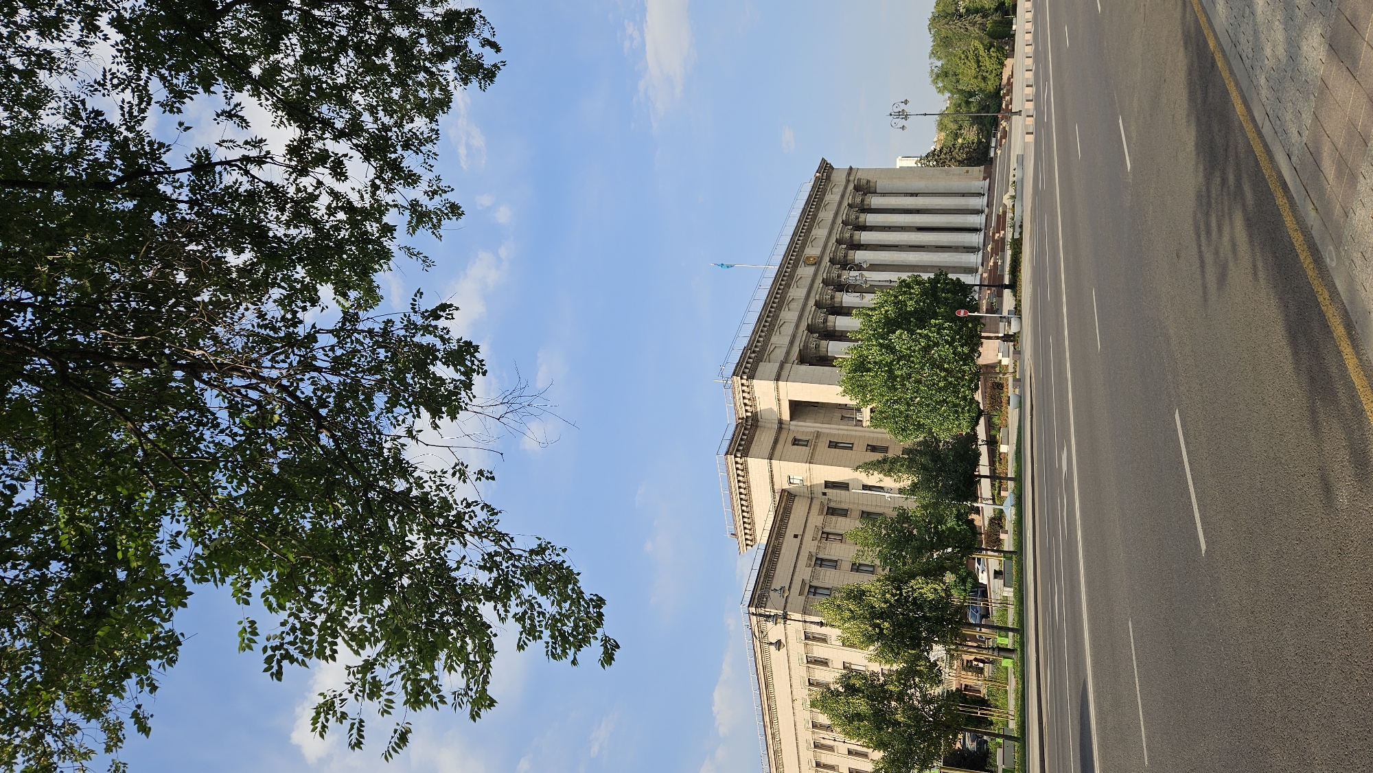 A wide asphalt road stretches across the foreground, marked with white lane dividers and a yellow curb. On the left side of the road, a row of green trees lines a sidewalk, with a large, imposing light-colored building with classical columns visible behind them. The building has multiple floors and numerous windows. A blue and yellow flag flies from the roof of the building. Lush green trees are also visible in front of the building and to its right. Overhanging the scene from the top of the image are the branches and leaves of a large tree, casting shadows on the sky. The sky is a clear blue with a few scattered white clouds. The overall scene depicts a street view of a prominent building in a city. This building is the Kazakhstan Hotel in Almaty, Kazakhstan.