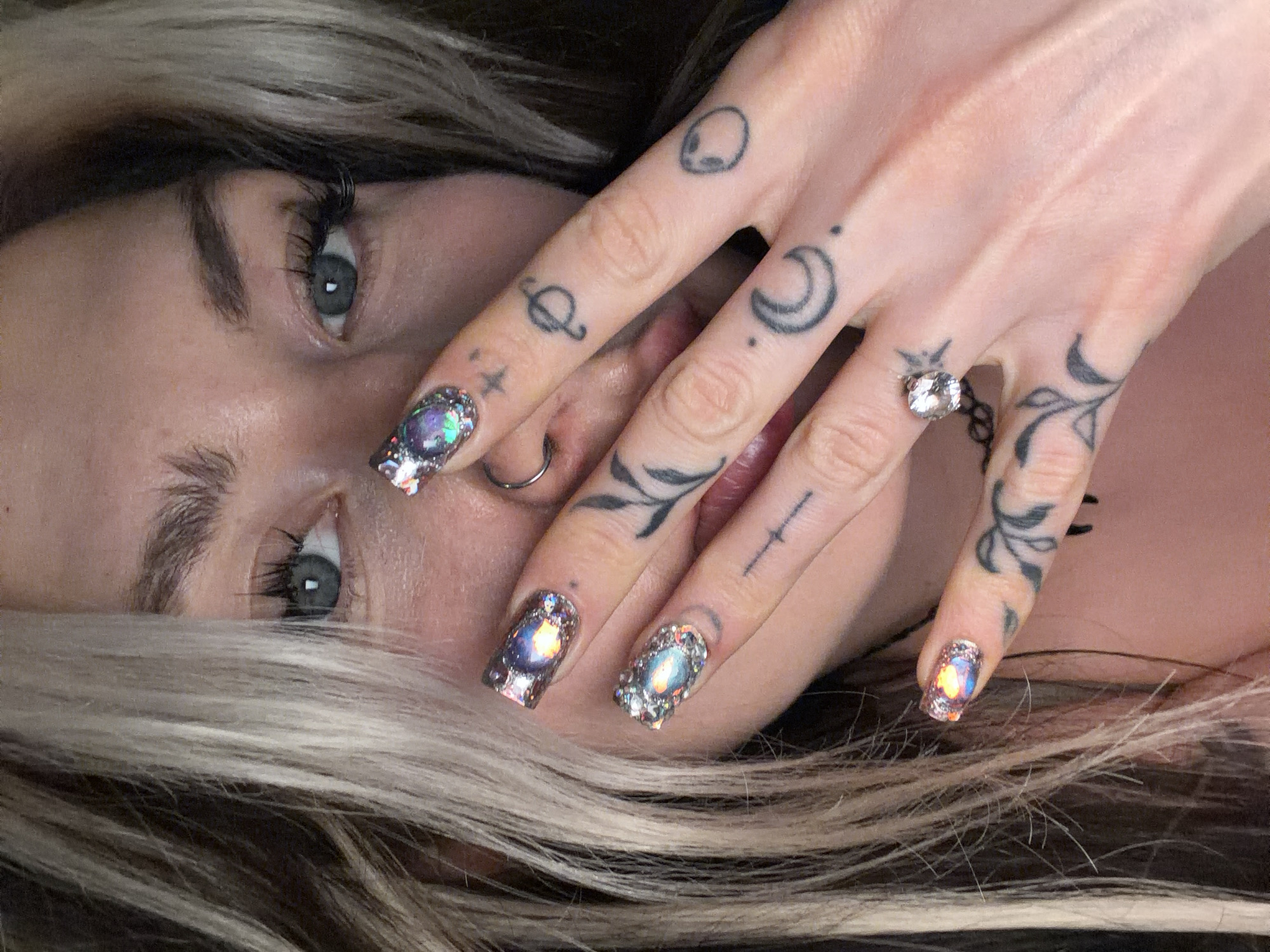 A close-up portrait features a woman with light skin, light grey eyes, and long, ashy blonde hair. The woman’s right hand is positioned near her face, partially obscuring it. Her fingers are adorned with small, black tattoos depicting an alien head, a planet, a crescent moon, a star, a cross, and leafy vines. A ring with a large, clear gemstone sits on her ring finger. Her nails are decorated with shimmering, iridescent nail art. A silver nose ring is visible in her left nostril.
The image does not provide any information about the location in the city of Budva, Montenegro.