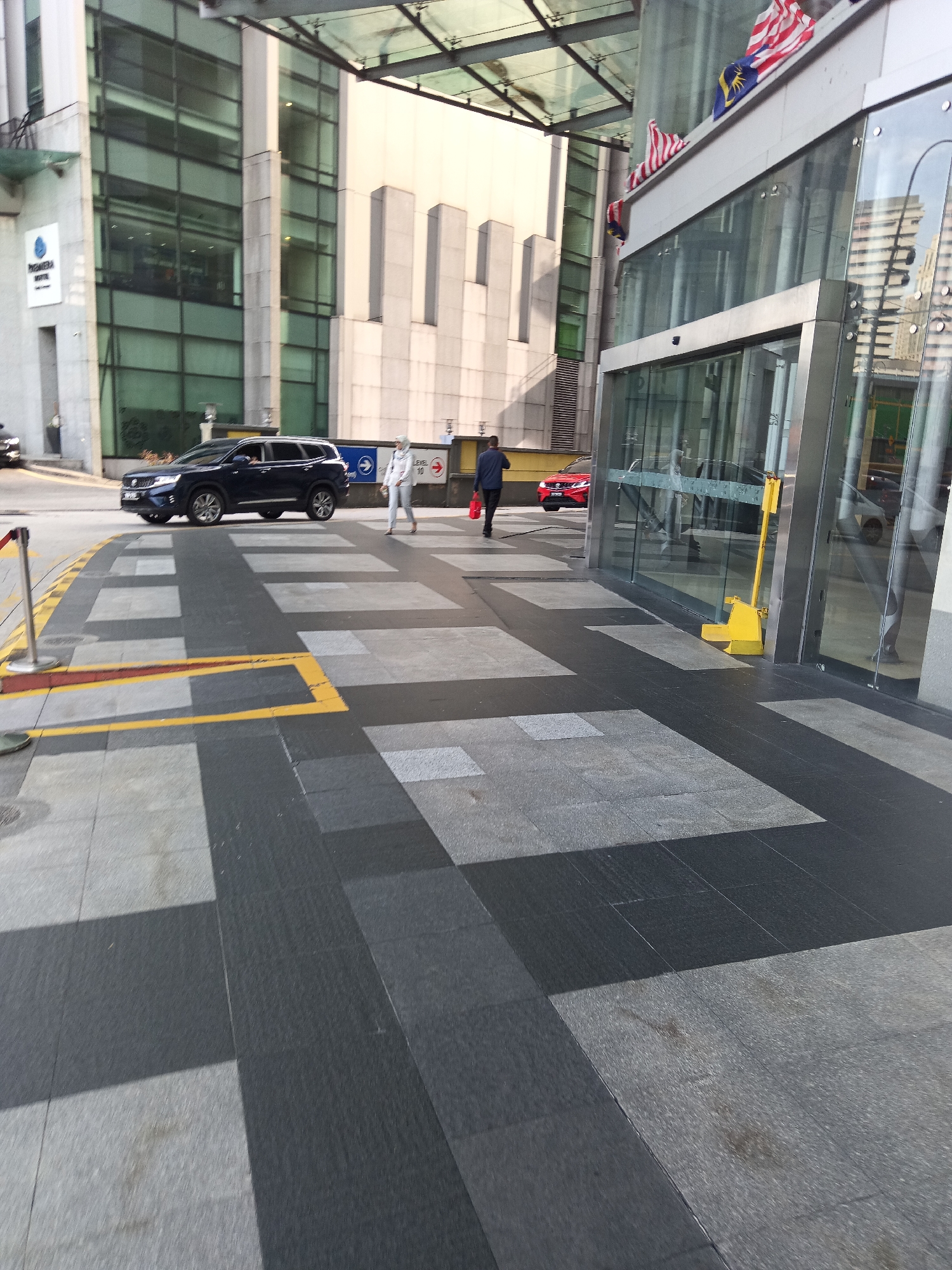 The image depicts the exterior of a modern building, likely a hotel, in Kuala Lumpur, Malaysia. The building features large glass facades and is adorned with Malaysian flags. In the foreground, a dark-colored SUV is parked near the entrance, while two individuals are walking across a pedestrian crossing. One person, dressed in white protective gear, is walking towards the SUV, and another person in dark clothing is walking away from the building, carrying a red bag. The ground is paved with a geometric pattern of dark and light gray tiles, with yellow markings indicating pedestrian lanes. The overall scene suggests an urban environment with ongoing activity.
