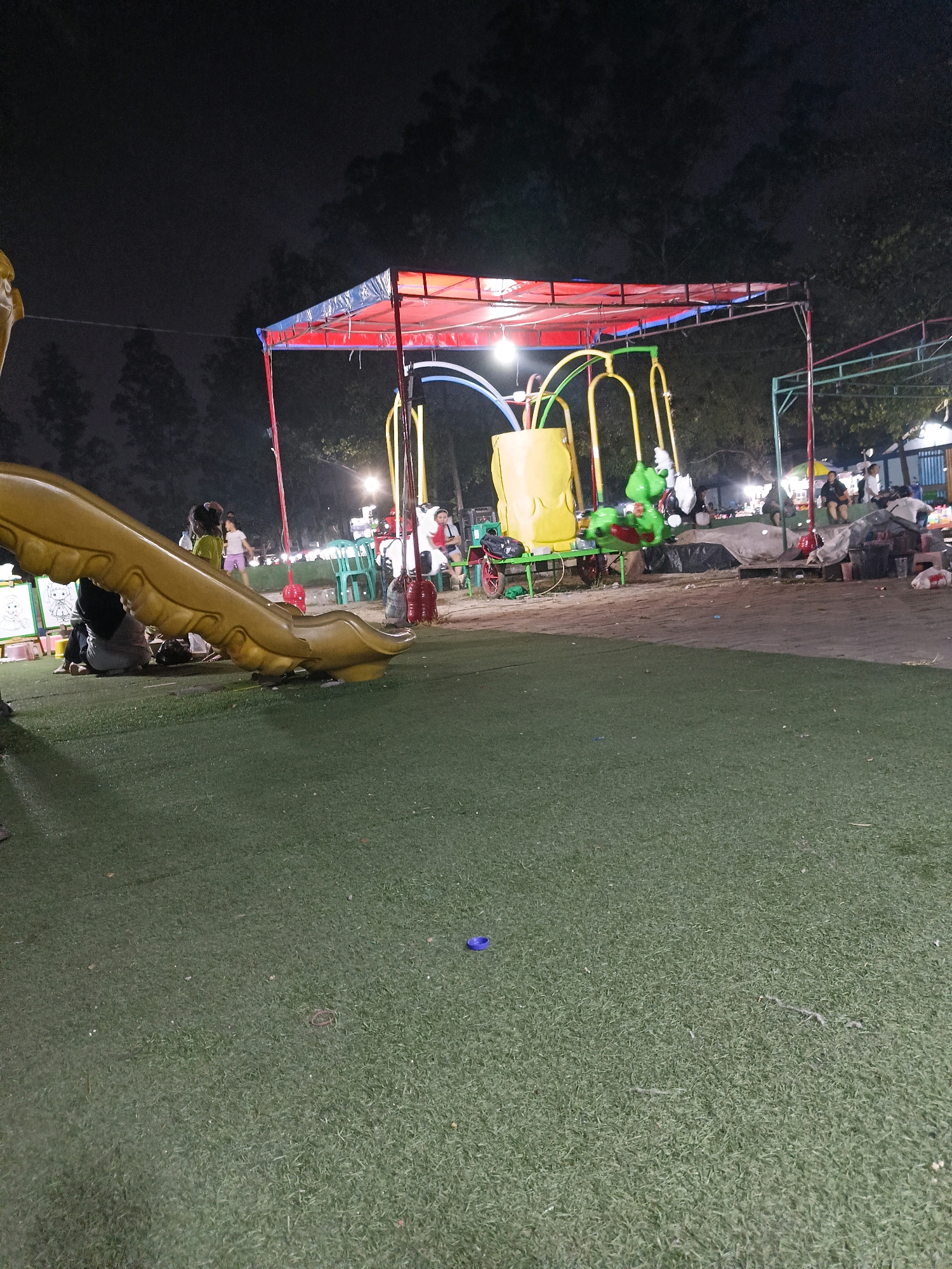 The image captures a nighttime scene at a playground in Tangerang, Indonesia. In the foreground, artificial green turf covers the ground. To the left, a large, yellow plastic slide curves upwards and is partially visible. In the mid-ground, a colorful playground structure with red and blue tarp roofing is set up. Yellow and green metal poles support the structure. A yellow bucket-like component of the playground is visible, along with a green dragon-shaped swing with a red seat, which is in motion. Several people are present in the background, including children and adults. Some are sitting on chairs, while others appear to be walking or standing near food stalls or vendors. Lights illuminate the area, casting a warm glow. Trees and foliage form a dark backdrop against the night sky. The overall environment suggests a lively, community-oriented recreational space.