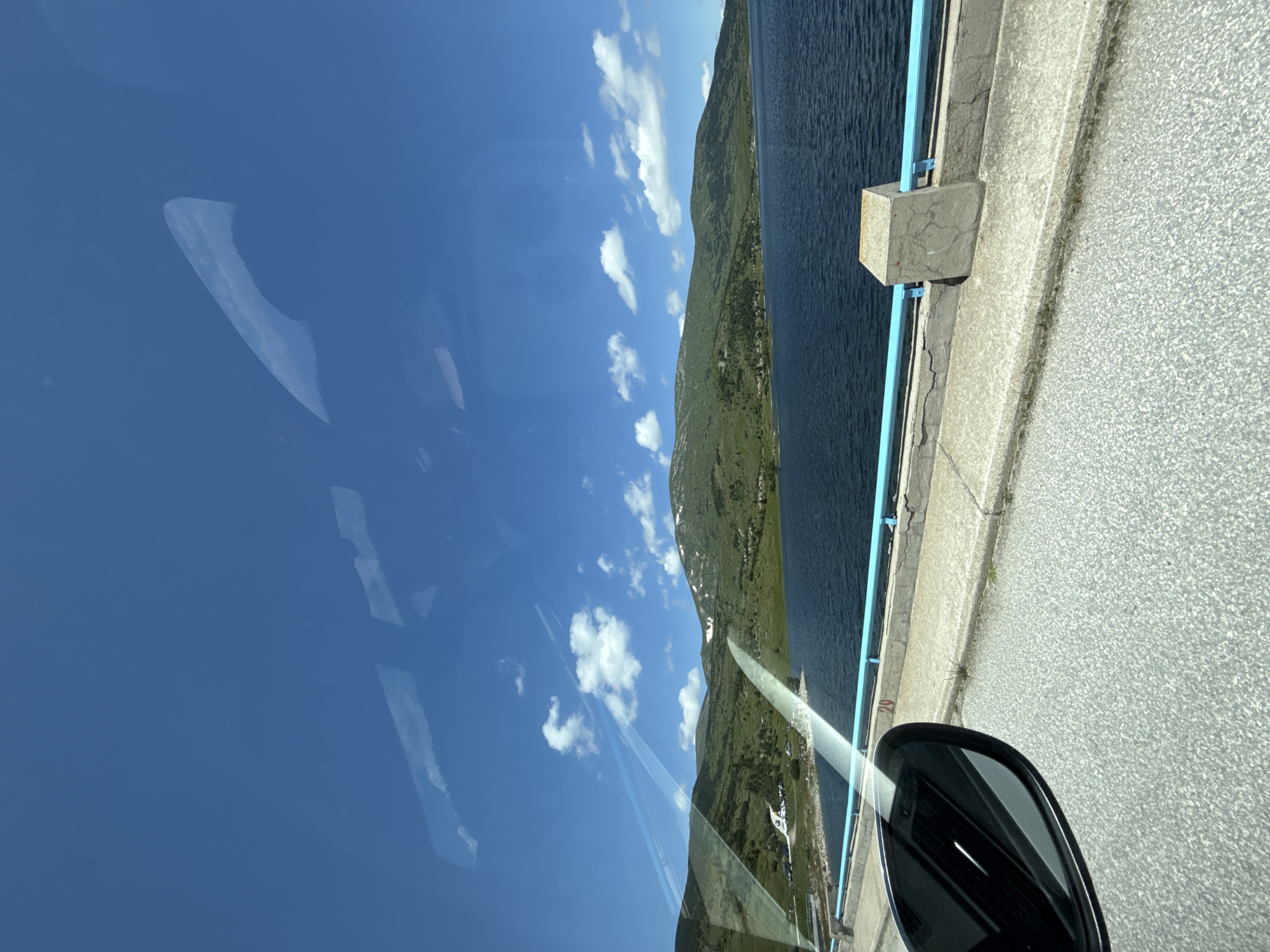 The image shows a wide shot of a lake on a sunny day, taken from a vehicle. In the foreground, part of a car's side mirror is visible on the left, reflecting the sky and clouds. The road surface is asphalt with visible aggregate. A concrete structure with a light blue railing runs along the edge of a dam or embankment. Beyond the railing, a large, dark blue body of water stretches across the frame. In the background, rolling green hills rise, some with patches of snow on their peaks. The sky is a vibrant blue with scattered white cumulus clouds. A small cluster of buildings and some parked vehicles are visible on the far side of the lake, nestled among the hills. The scene appears to be of the Georgi Dimitrov Dam, located near the village of Smolevo, Bulgaria, a common tourist and recreational area. There are no people visible in the image.