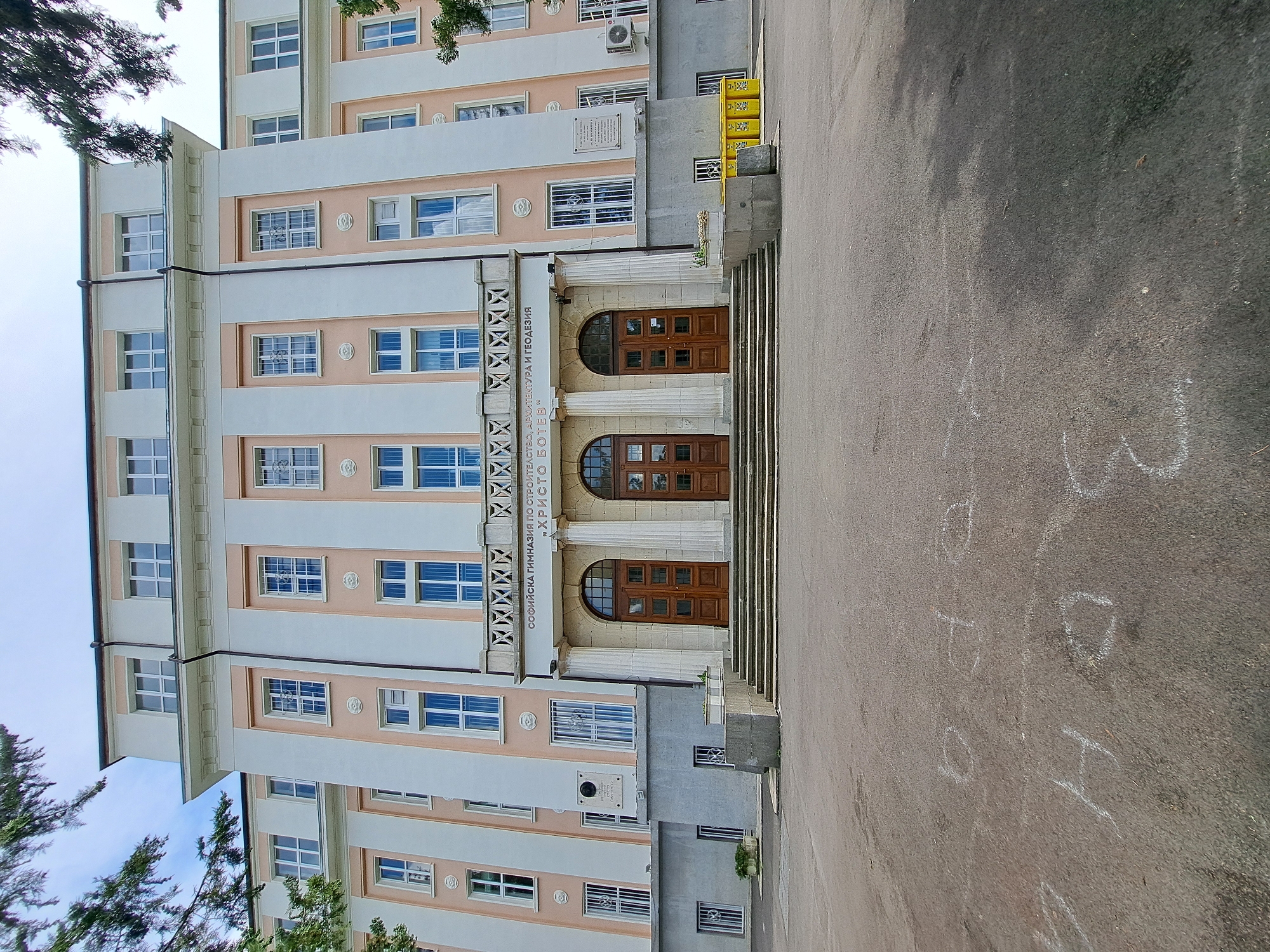 The image is an exterior shot of a building, likely a school, with an entrance featuring large wooden doors and classical columns. The building is multi-story with many windows and decorative architectural elements, painted in shades of cream and light pink. To the right of the entrance, there is a paved area with chalk drawings on the ground. The scene is captured during daylight, with the sky visible at the top left and some trees bordering the building on the left. A plaque is visible on the building facade above the entrance. The inscription on the building above the entrance reads "СОФИЙСКА ГИМНАЗИЯ ПО СТРОИТЕЛСТВО И АРХИТЕКТУРА „ХРИСТО БОТЕВ“" which translates to "SOFIA HIGH SCHOOL OF CONSTRUCTION AND ARCHITECTURE 'HRISTO BOTEV'". The chalk drawings on the ground are illegible. The image is rotated 90 degrees counter-clockwise.
