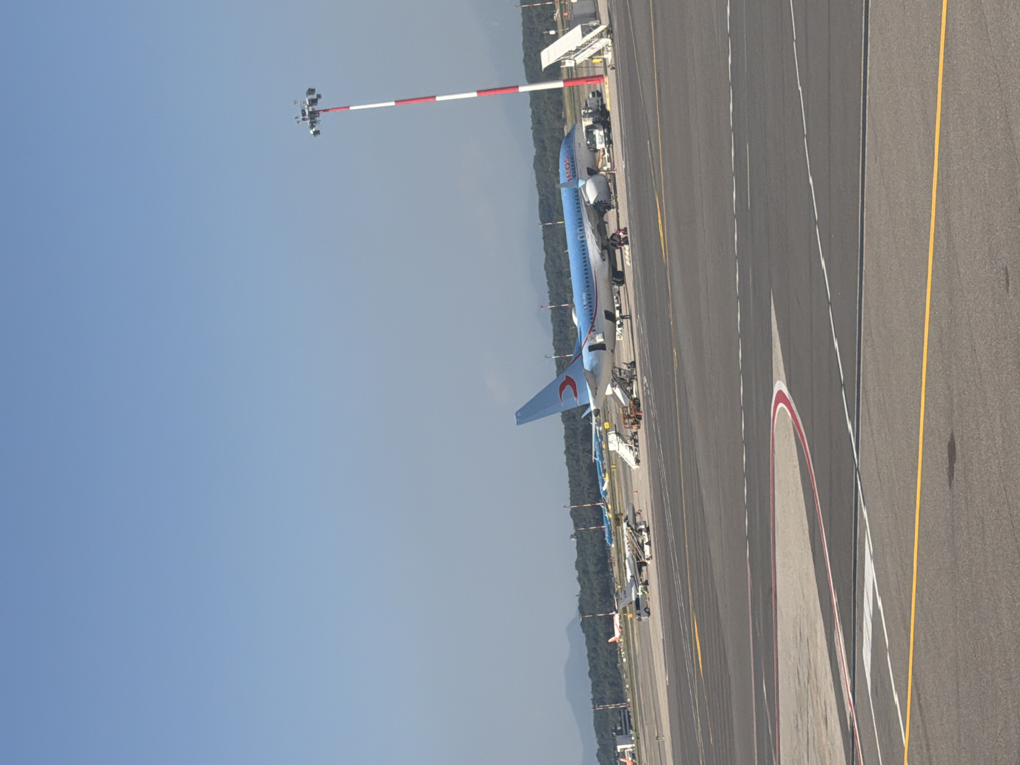 This image, taken at Malpensa Airport in Ferno, Italy, shows an airport tarmac on a clear day. In the foreground, the asphalt of the runway is visible with yellow and white markings. In the midground, several commercial aircraft are parked. A large blue and white passenger jet with the TUI logo is prominently featured, attended by ground crew and service vehicles. Smaller aircraft, including a light gray jet with a red stripe, are also present. In the background, a dense line of trees forms the horizon, with faint mountains visible beyond. A tall, red and white striped light pole stands near the blue and white aircraft. The sky is a clear, pale blue.