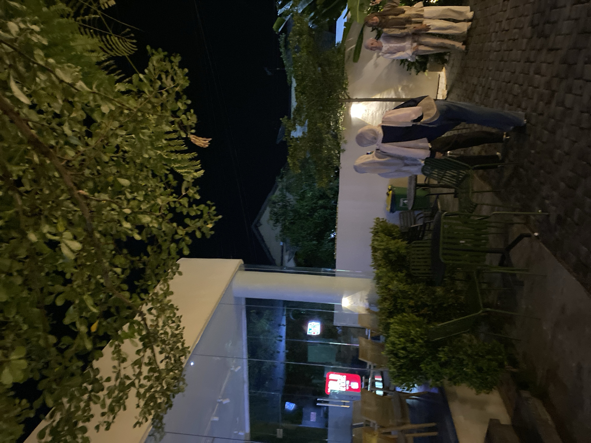 This media file depicts an outdoor scene at night, likely a cafe or restaurant patio in Lhokseumawe, Indonesia. The dark sky is illuminated by various artificial lights, creating a tranquil evening atmosphere.
In the foreground, a person wearing a light-colored jacket or hoodie and blue jeans is seen bending forward. Further back and to the right, two static figures resembling mannequins or statues, dressed in flowing, light-colored attire, stand on a dark, textured paved surface. Another individual is partially visible behind the bending person.
The scene features green outdoor tables and chairs, suggesting a dining or social area, with a green trash bin placed near the seating. To the left, a modern building with extensive glass walls reflects interior lights and glowing signs. One notable red sign with indistinct white text is visible through the glass. Lush green trees and foliage frame the scene, growing prominently on the left side, along the building, and in the background. The ground beneath the seating area is composed of dark paving stones.