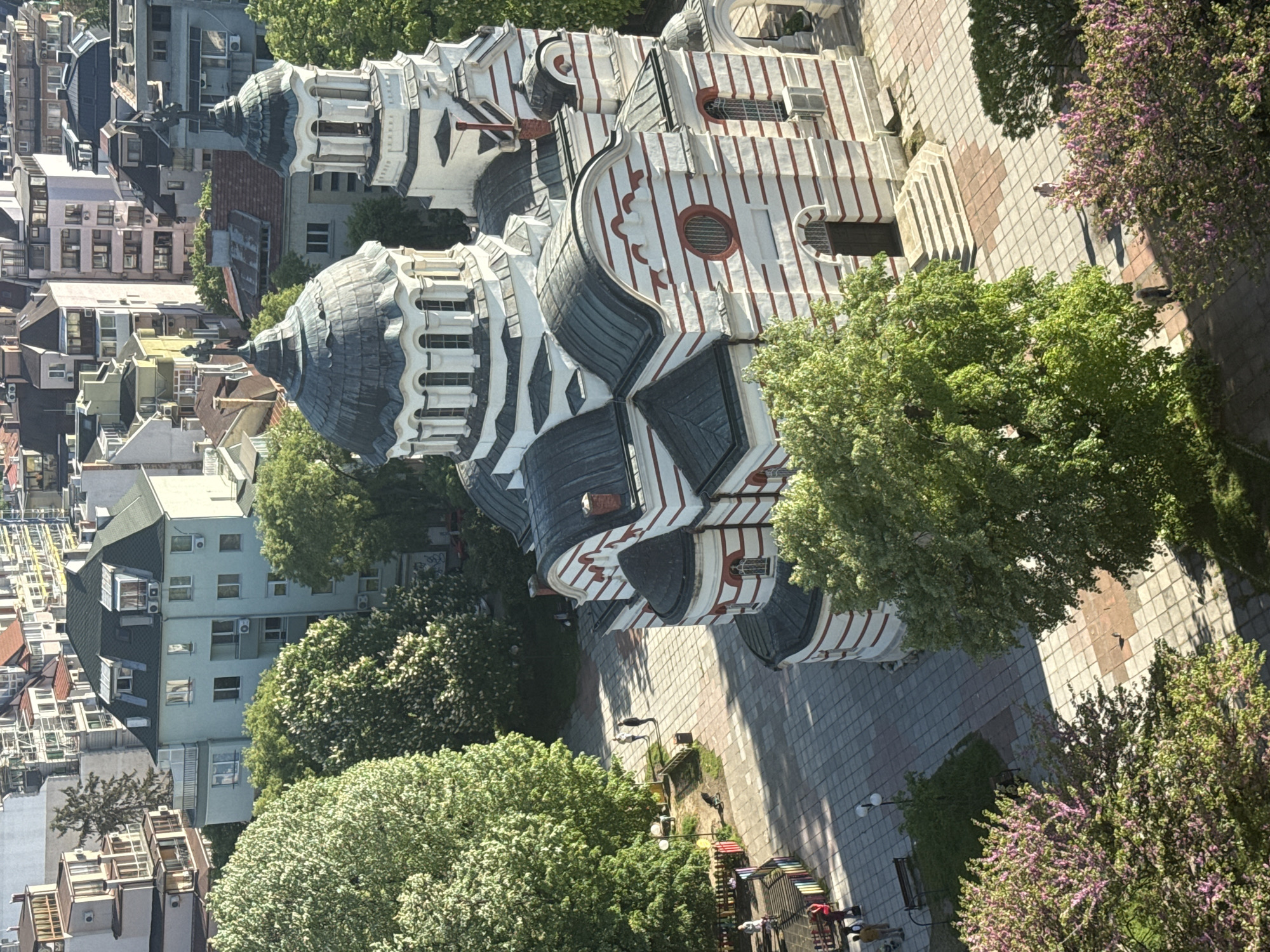 The image shows an aerial view of the Dormition of the Mother of God Cathedral in Varna, Bulgaria. The cathedral is recognizable by its white facade with red horizontal stripes, dark gray domes, and ornamental details. The surrounding area includes buildings, trees, and a pedestrian walkway. The pedestrian area has brick pavers and benches and streetlights. There are several flowering trees with purple flowers. Buildings of varying heights and architectural styles are seen surrounding the cathedral and green trees dot the scene. The environment appears to be urban, during daytime.