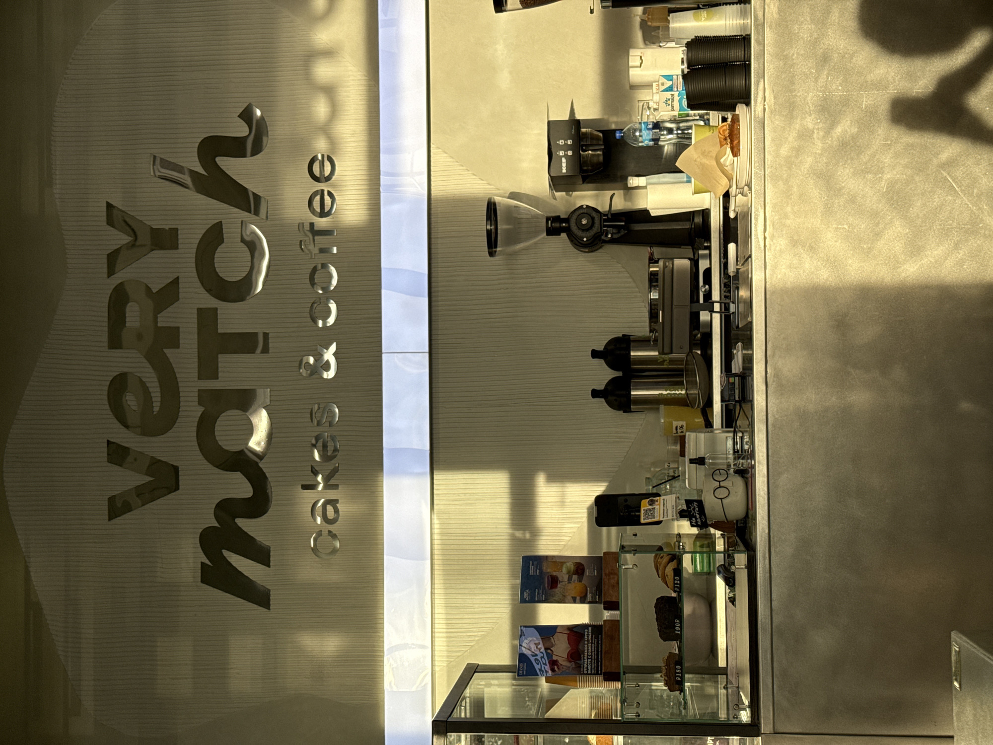 This is a vertical, eye-level shot of a cafe interior in Nizhny Novgorod, Russia. The cafe features a metallic, striped wall on the left with large, embossed text that reads "very match cakes & coffee." To the right of the wall, a counter area is visible, displaying various coffee-making equipment, including a grinder and espresso machine. There are also bottled beverages, cups, and shelves stocked with snacks and pastries. A glass display case in the foreground shows several cakes and baked goods, some with price tags. Sunlight streams in from the right, casting shadows on the counter and wall, suggesting it might be late afternoon. The overall atmosphere appears to be that of a functional cafe ready for business. The visible text includes the cafe's name and prices such as "P120", "190P", and "P180".