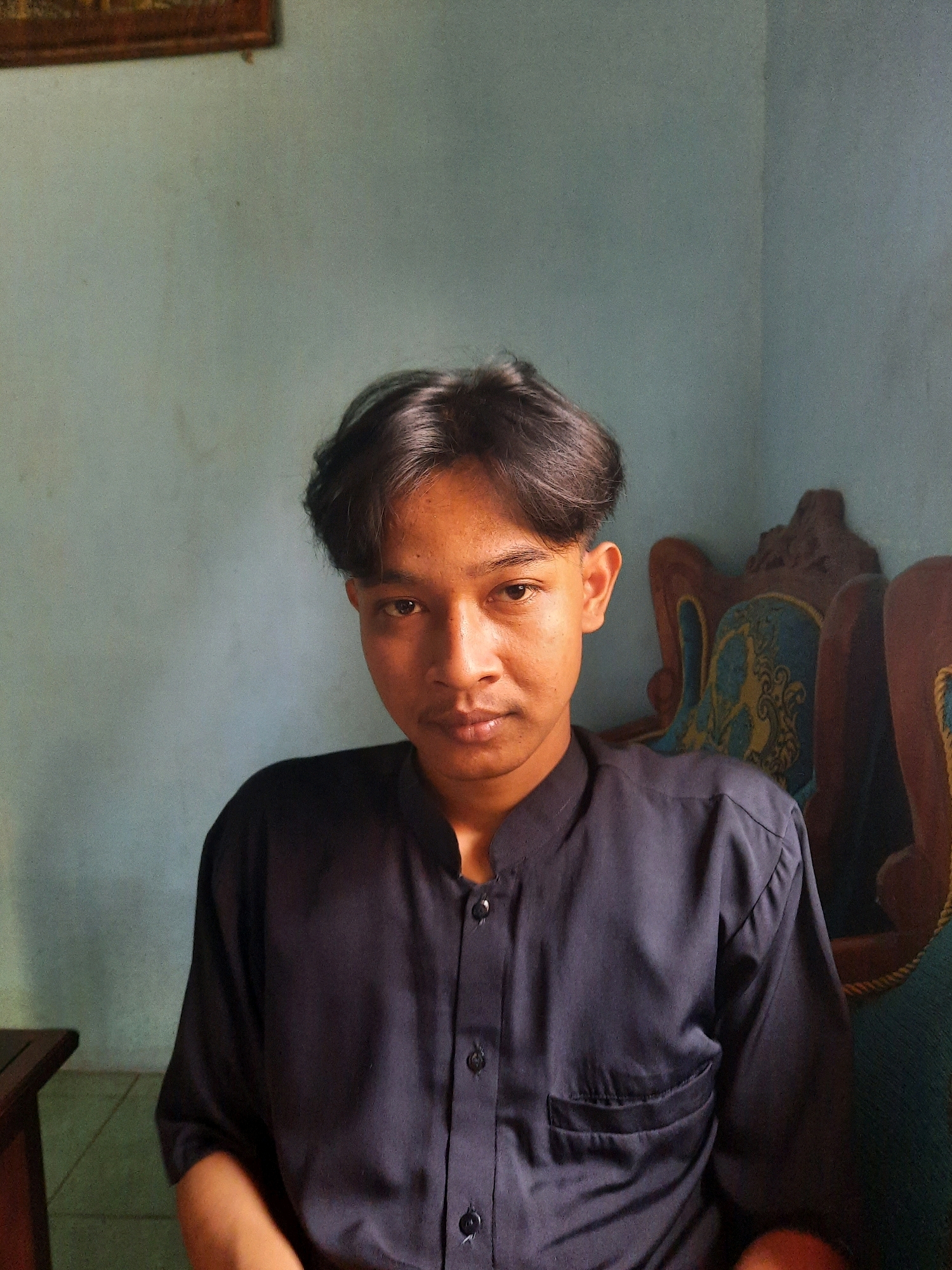 The image shows a young man with dark, wavy hair and brown eyes, looking directly at the camera. He is wearing a dark blue, collared shirt with buttons down the front and a chest pocket. The background consists of a light blue wall with a painting on the upper left and decorative chairs with carved wooden frames and patterned upholstery behind him. The lighting is soft, suggesting an indoor setting, possibly in a home. Based on the visual cues, the location appears to be Suren, Indonesia, a common type of interior setting in residential areas of the country.