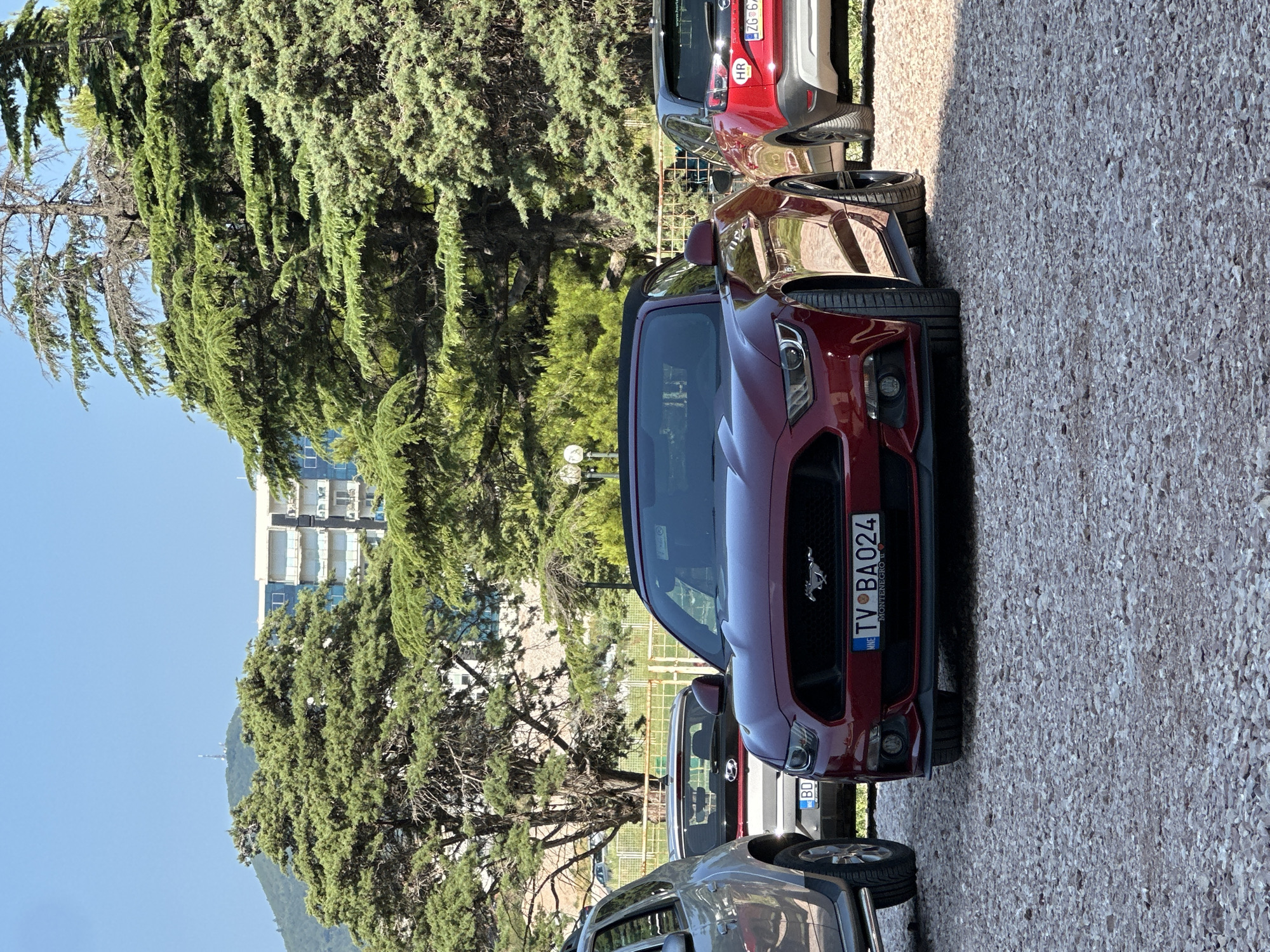 In Boreti, Montenegro, under a clear blue sky, a dark red Ford Mustang convertible is parked on a gravel lot. The car is positioned centrally in the frame, facing the viewer. To the left of the Mustang, a portion of a silver SUV is visible, and behind it, a white Hyundai is parked. To the right of the Mustang, a red Opel is parked. The background features lush green trees and a modern multi-story building with balconies, with a distant mountain topped with communication towers. The scene suggests a sunny day in a coastal or resort area.