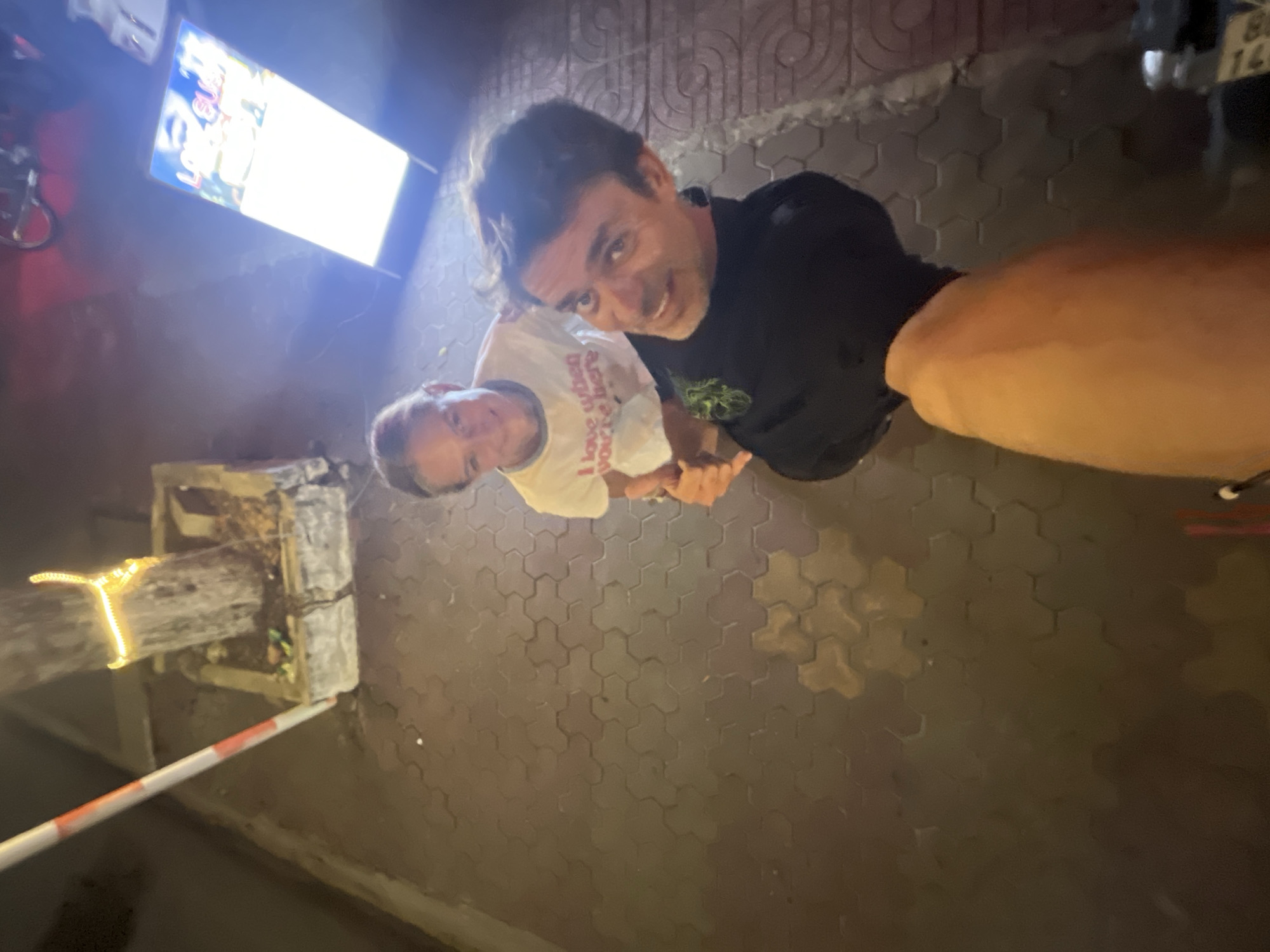 In the city of Phu Thuy Ward, Vietnam, at night, a low-angle selfie is captured on a sidewalk paved with hexagonal bricks. Two individuals, a young female and a male, are the primary subjects. The young female, positioned behind the male, is wearing a white t-shirt with red lettering that reads "i love when you're here". She has her hair pulled back and is smiling towards the camera. The male, in the foreground, is wearing a black shirt with a green emblem on the left chest. He is looking directly at the camera and appears to be giving a thumbs-up gesture. His right arm is extended upwards, holding what appears to be the camera or phone, with his forearm visible in the bottom of the frame.
Behind the subjects, a tree trunk is visible, decorated with a string of yellow lights. The tree is situated within a concrete planter box. To the left of the tree, a white and orange striped pole stands upright. In the background, a bright, illuminated display board is positioned on the right side, showing text and images. The overall environment suggests an outdoor urban setting with artificial lighting.