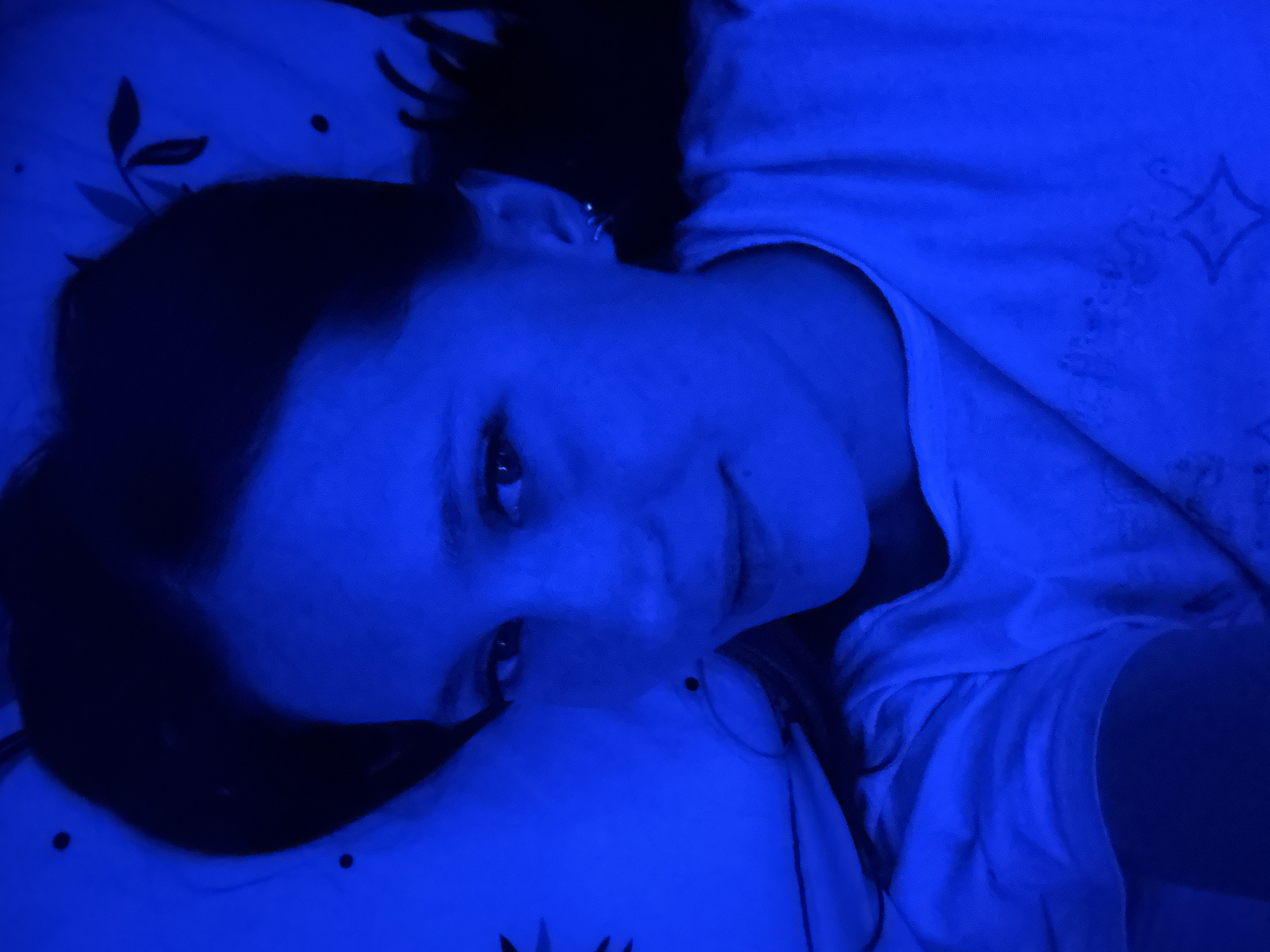 A person lies on a pillow with a patterned design, illuminated by blue light. The location cannot be determined to be Chaikovsky, Russia, from the image alone. The person is wearing a white t-shirt.