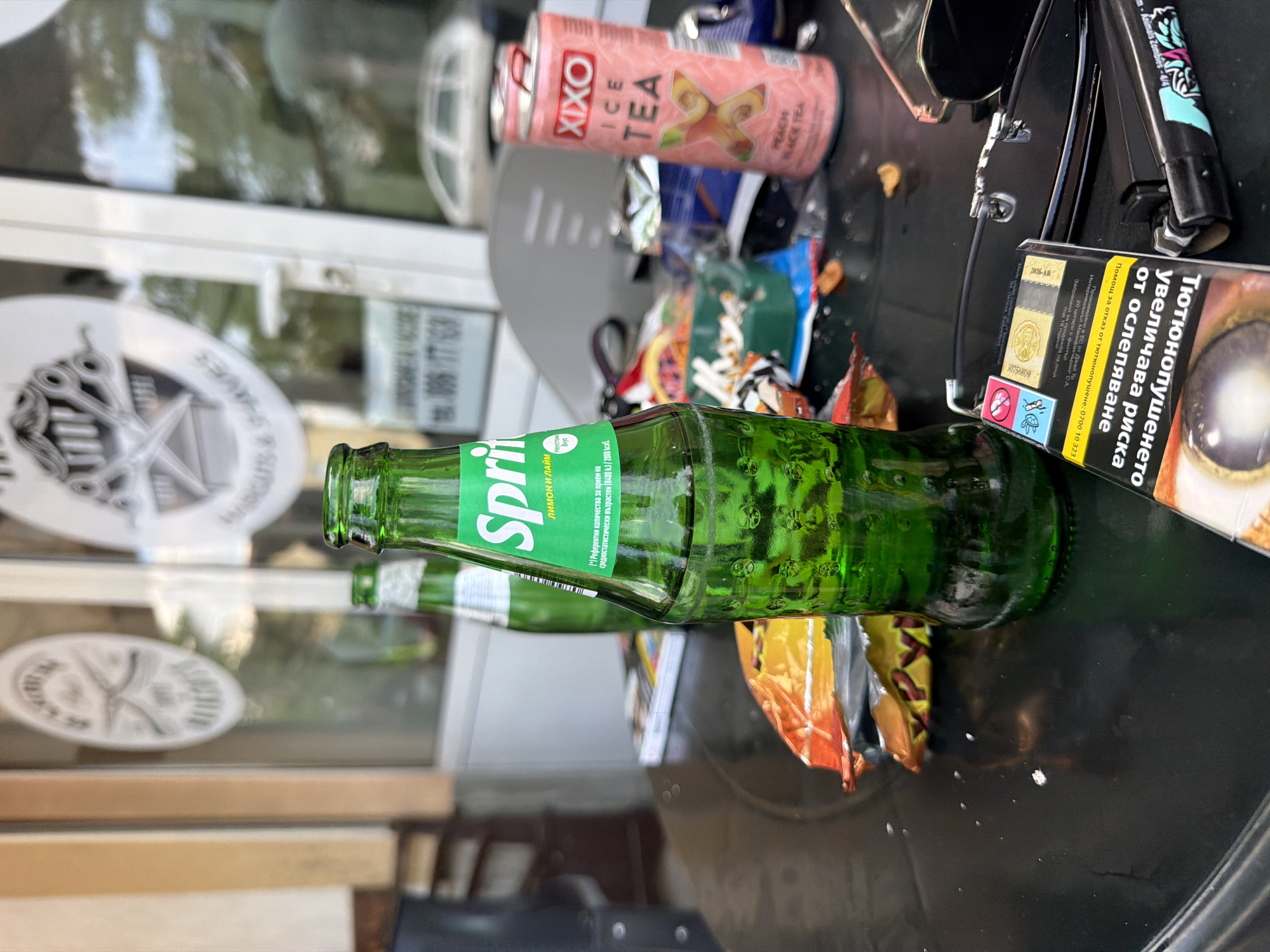 This image captures a cluttered outdoor table scene outside a commercial establishment, specifically identified as a barber shop based on window decals, located in Dabene, Bulgaria.
In the foreground, a black, glossy outdoor table is covered with various items indicating recent use. A prominent green glass bottle of Sprite, partially consumed, features a Bulgarian label stating "Sprite," "Лимон и Лайм" (Lemon and Lime), and nutritional details. To its right, a pack of cigarettes is present, displaying a Bulgarian health warning: "Тютюнопушенето увеличава риска от ослепяване" (Smoking increases the risk of blindness). Adjacent to the cigarette pack are a black lighter with some graphic design and a pair of dark sunglasses, partially folded.
Further on the table, an open, crumpled orange and yellow snack bag is visible, along with a pink Xixo Ice Tea can, labeled "Peach Black Tea." A small, green ashtray containing multiple cigarette butts indicates smoking activity. The table surface is marked by crumbs and other small debris, suggesting a used and untidy state. Another green bottle, similar in shape to the Sprite bottle, is partially visible further back on the table.
The background consists of a glass door or large window reflecting the surroundings. Visible through and reflected on the glass are blurred trees or foliage and what appears to be a white vehicle. Two white vinyl decals are affixed to the glass: one is a circular logo depicting crossed scissors and a razor, with text partially legible as "BARBERS, CUTS & SHAVES." Below this, another decal displays a phone number: "006 177 163." Faint reflections of indistinct figures or objects are present within the glass. The interior of the establishment, seen through the glass, shows light-colored walls and general ambient lighting. No individuals are clearly identifiable in the scene.