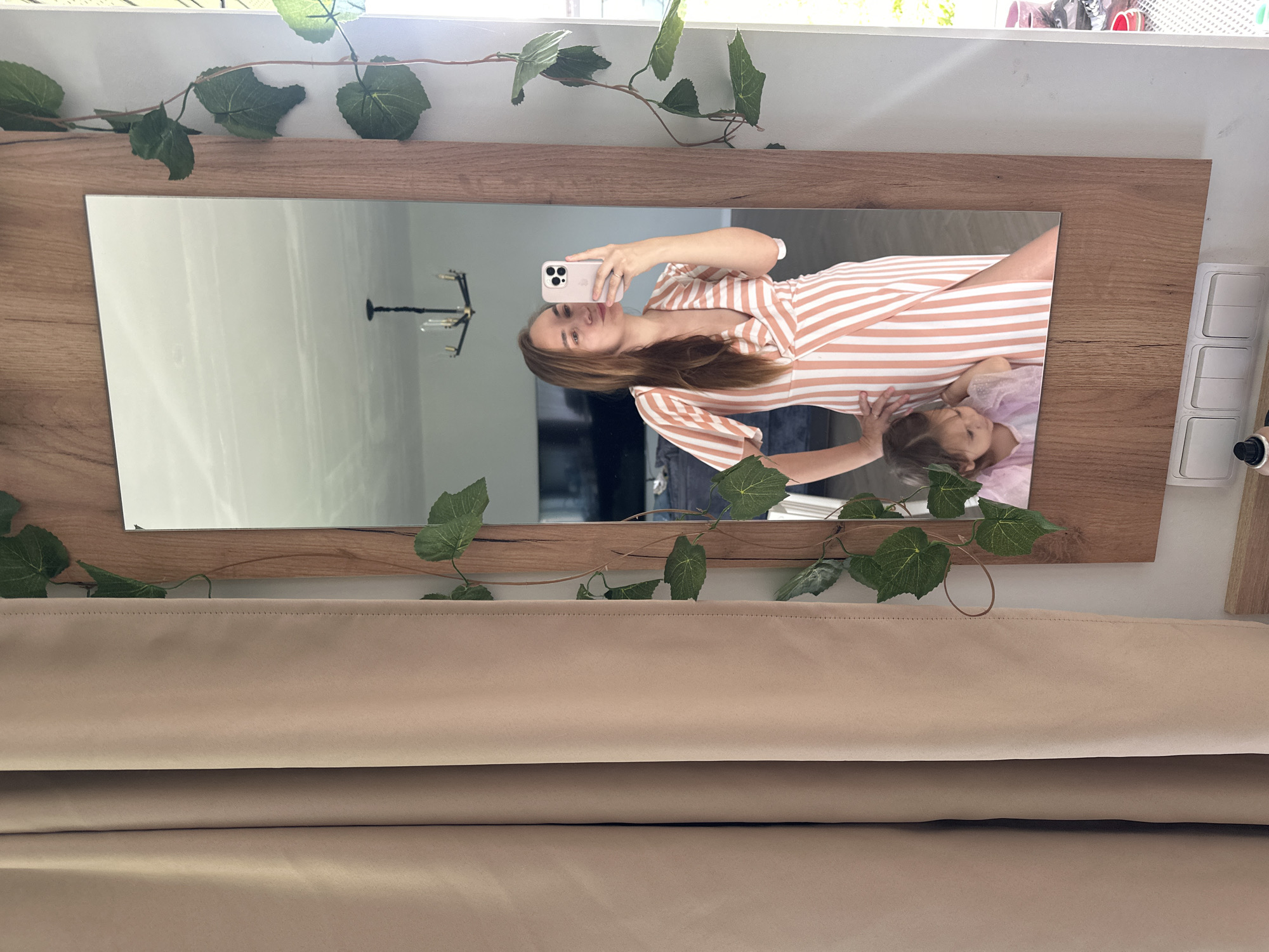 A woman is taking a mirror selfie in what appears to be a residential interior, possibly in the city of Kharnavy, Russia. She is wearing a peach and white striped wrap dress with short sleeves and a plunging neckline. Her hair is long and brown, and she is holding a pink smartphone in her right hand. Her left hand is on her hip. Reflected in the mirror behind her is a young girl wearing a pink dress. The mirror is framed with a wooden border and decorated with artificial green ivy. To the left of the mirror is a beige curtain. In the reflection of the mirror, a chandelier is visible on the ceiling, and a television is to the left. Below the mirror, there are three white light switches on the wall.